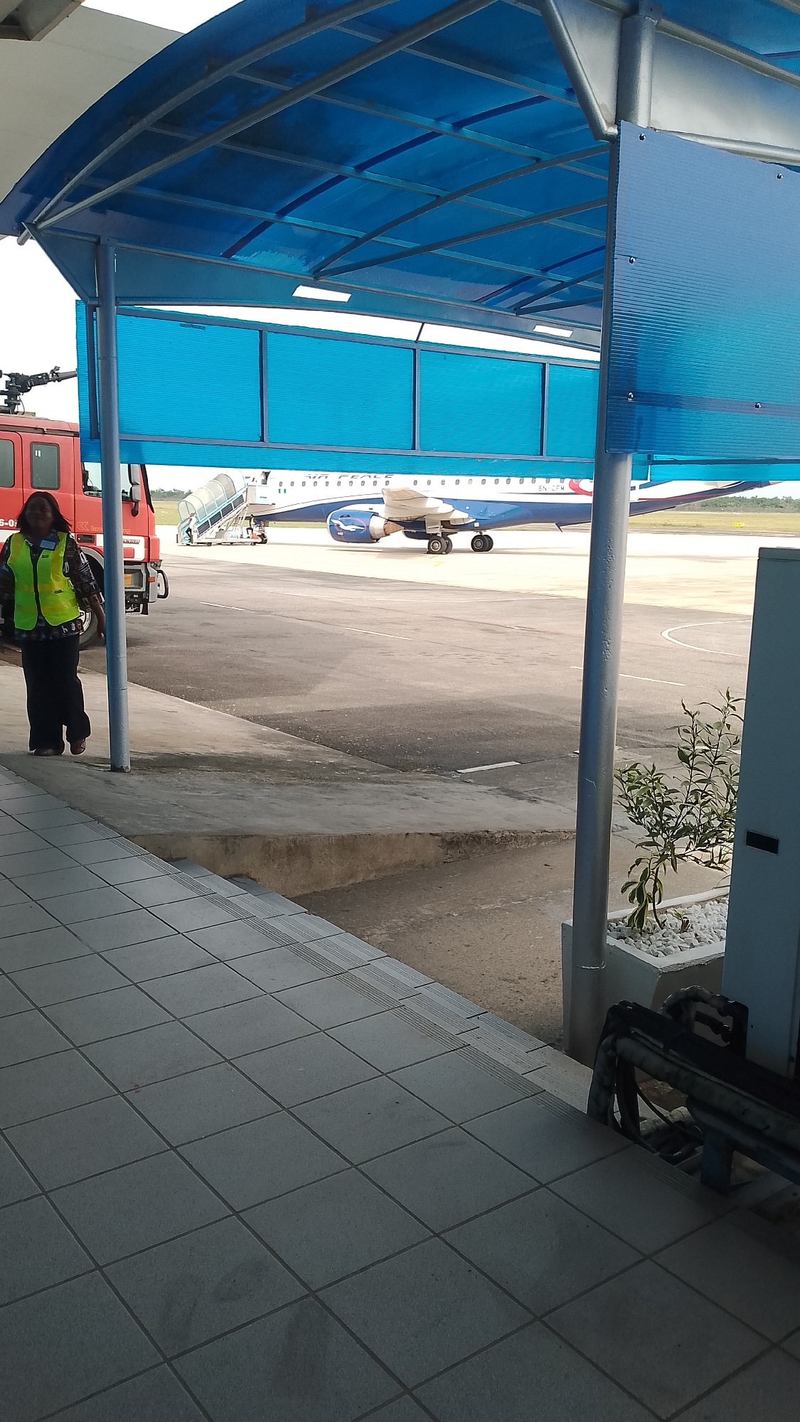 The image shows an airport scene in Orerokpe, Nigeria. A woman wearing a neon green vest and dark clothing is walking left to right on a tiled walkway. Behind her, on the tarmac, there's a red fire engine and an airplane, presumably an Air Peace jet, with a boarding stairwell leading up to the cabin. A blue metal structure with a blue translucent roof and side paneling covers part of the walkway. A small plant in a white container is also visible, along with metal railing near the right edge of the scene.