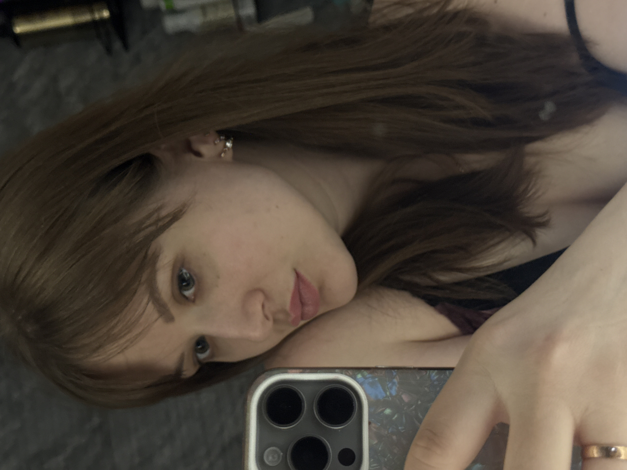 A fair-skinned person with light brown hair takes a selfie. They have green eyes, light pink lips, and an earring in their left ear. A smartphone is visible in their right hand, with a case featuring a speckled design. A simple gold ring is on their ring finger. The background appears to be a gray surface, possibly a bed or floor. No other identifying information relating to the city of Chaikovsky, Russia, is visible.