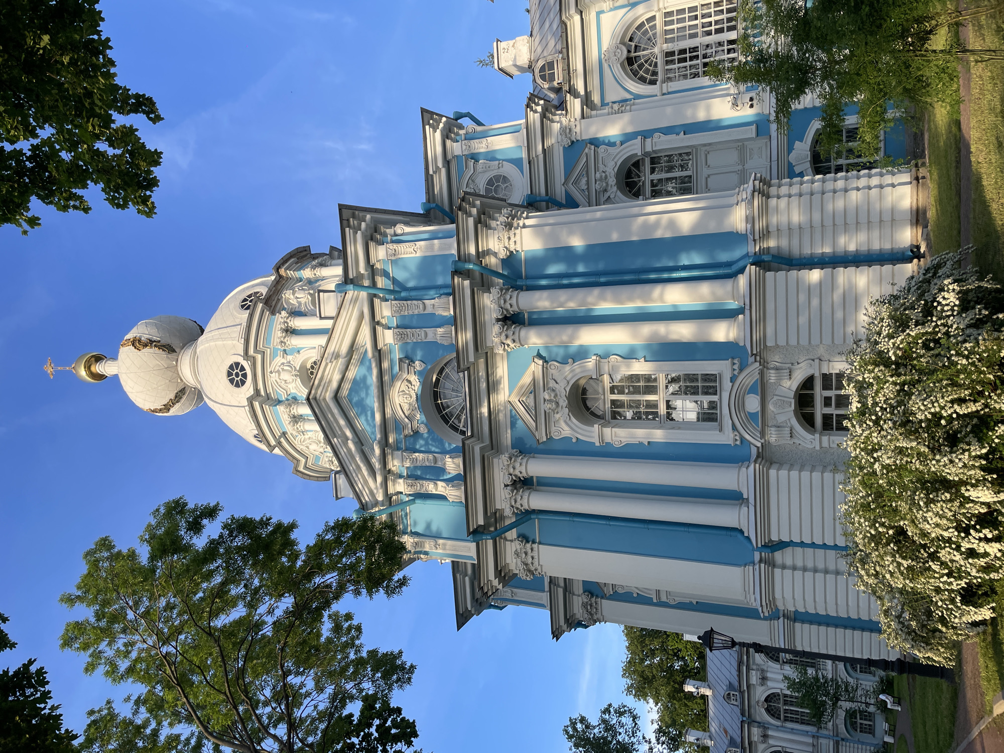 A low-angle, exterior view captures the Smolny Cathedral in St. Petersburg, Russia, under a clear blue sky, illuminated by late afternoon sunlight. The cathedral, a prominent example of Russian Baroque architecture, features a distinctive light blue and white facade. Numerous white Corinthian columns, decorative white stucco work, and ornate arched windows with intricate grilles adorn its multi-tiered structure. A large white central dome, topped with an onion-shaped finial and a golden cross, dominates the upper portion of the building. Blue rain gutters are visible on the facade, running vertically.
Green deciduous trees with dense foliage partially frame the cathedral on the left and upper right. In the foreground, a large, dense bush with numerous small white flowers extends across the base of the building. A paved pathway is visible on the left, alongside a black street lamp. Further to the left, part of a less ornate, lighter-colored building with arched windows and a gray roof, belonging to the Smolny Convent complex, is visible. One person, dressed in white, is seated on a white bench under a tree in the mid-ground on the left. The lighting creates distinct shadows on the building's blue sections, cast by the white columns, and highlights the vegetation with a warm glow. The sky is a bright, clear blue with a few faint wisps of clouds.