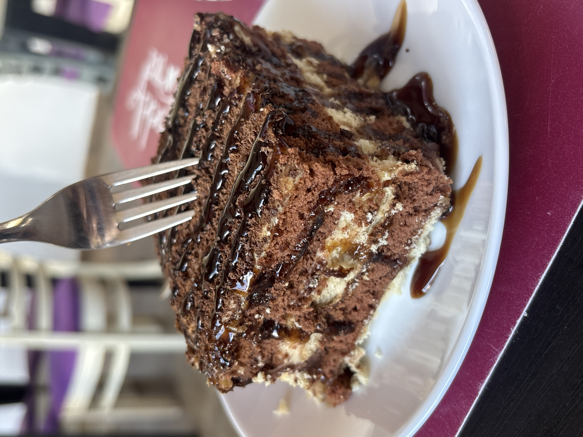 A close-up, slightly angled shot shows a slice of marbled chocolate cake on a white plate. The cake has layers of dark chocolate and lighter sponge, with a rich chocolate sauce drizzled over the top. A silver fork is positioned on the left, with its tines touching the cake. The plate is set on a dark surface, possibly a table, next to a burgundy placemat or tablecloth. In the background, out of focus, are elements of a cafe or restaurant setting, including what appears to be a pink paper item with some faint, elegant script, possibly a menu or a brand logo, and some blurred white furniture with purple accents. The lighting suggests it's daytime, and the overall mood is one of indulgence and enjoyment of a dessert. No specific emotional cues are evident from the image itself. The text in the background is too blurry to read. The location context is Chavdar, Bulgaria.
