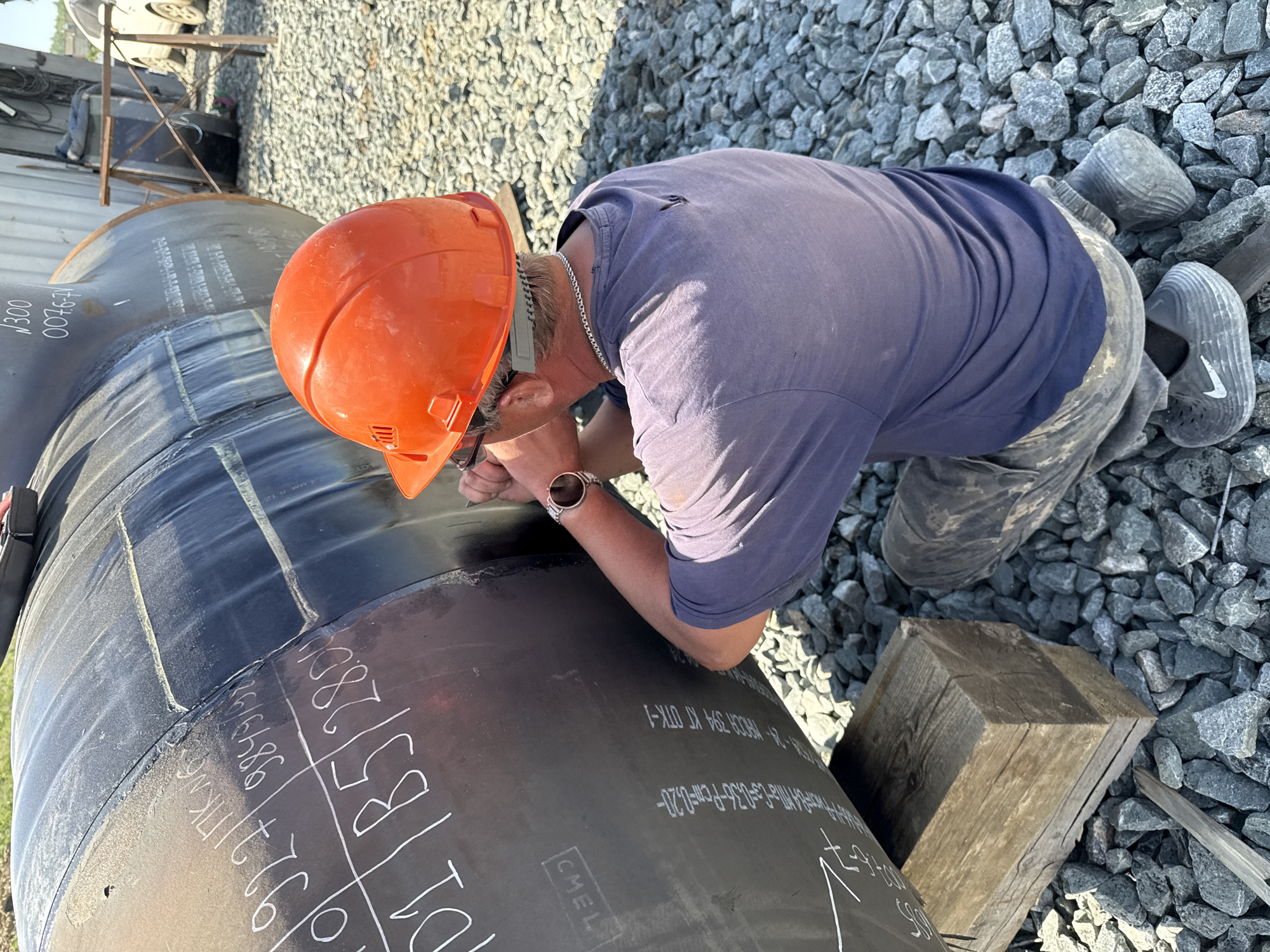 A man wearing an orange hard hat, a navy-blue T-shirt, and camouflage pants is bending over and inspecting a large, black pipeline lying on a bed of gray gravel. He has a silver chain around his neck and a watch on his left wrist. The pipeline has white markings and numbers written on it, including "927", "9849/117", and "01/B5/280". A wooden block is visible near the pipeline. The location is outdoors and appears to be a construction or industrial site. The scene shows signs of construction activity. The photograph does not provide specific information that identifies the location as Leninsk, Russia.