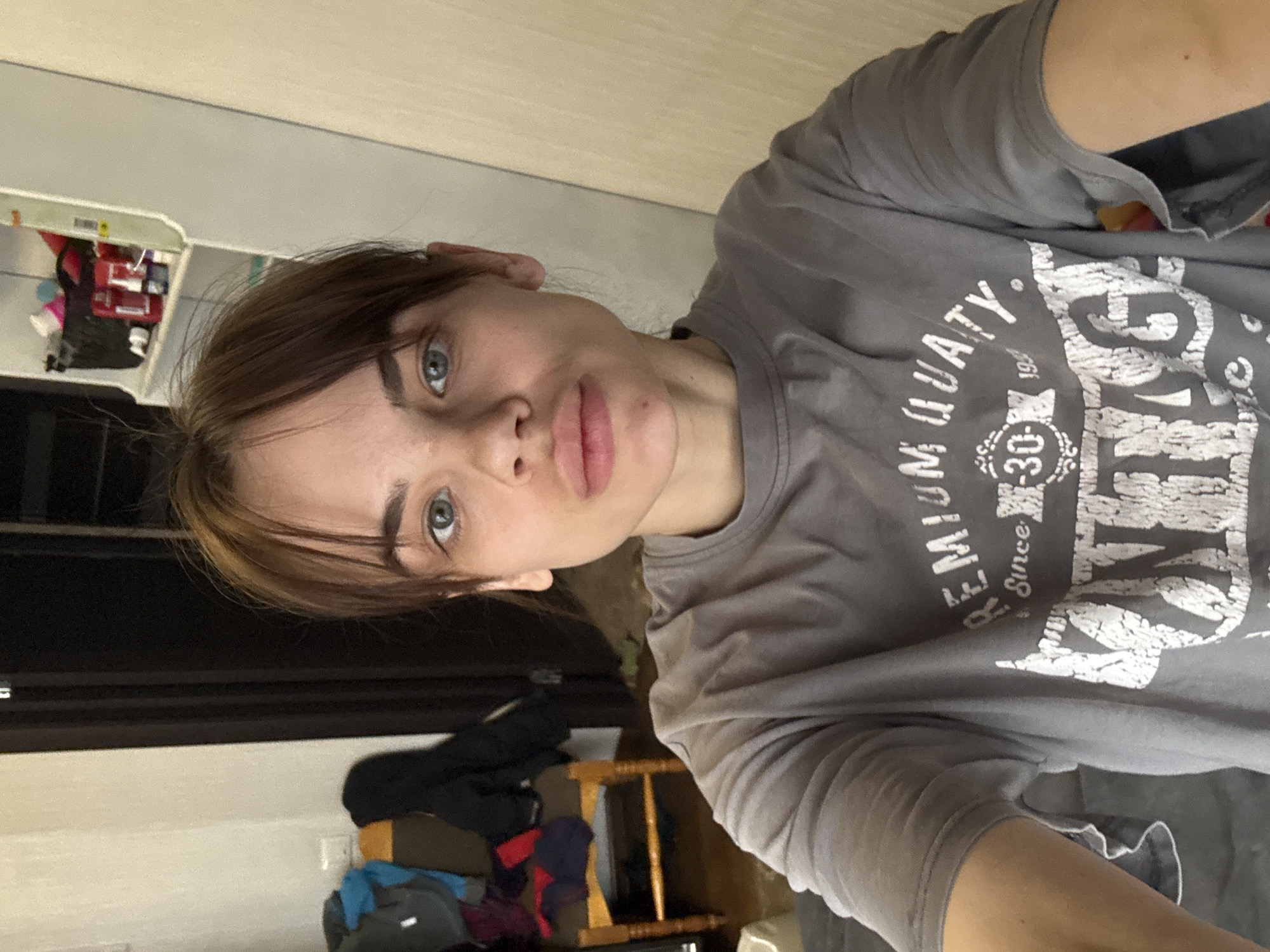 A selfie-style, indoor photograph shows a woman with fair skin, light brown hair, and blue eyes. She is wearing a gray t-shirt with white lettering that reads "PREMIUM QUATY Since 1930." The background shows a portion of a white wall, a dark door, and a shelf with miscellaneous items on it. The specific location within Rostov-on-Don, Russia, is not discernible from the visual information provided.