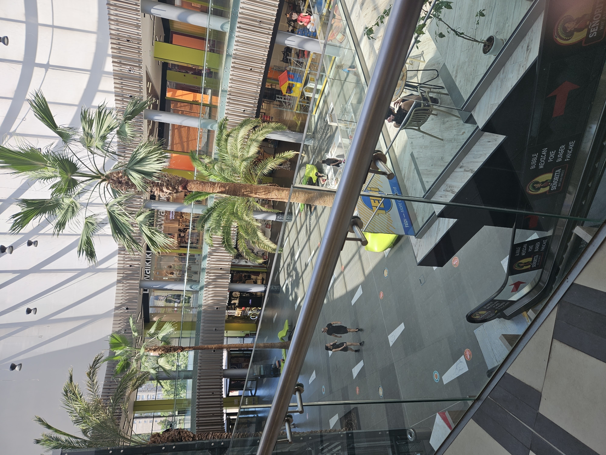 The image depicts the interior of a modern shopping mall, likely located in Stara Zagora, Bulgaria, given the context. The scene is viewed from an elevated position, looking down onto a multi-level shopping area with a contemporary architectural design. Palm trees are a prominent feature, adding a tropical element to the indoor environment. The floors are tiled in a dark gray and white pattern.
On the ground level, two individuals, a man and a woman, are walking across the floor. They are dressed in dark athletic wear. Scattered on the floor are white and circular markings, possibly indicating social distancing guidelines. A bright green, ergonomic bench is visible in the background. Further in the distance, a children's play area is set up with colorful structures. Shops with brightly colored facades, featuring orange, yellow, and green accents, line the upper levels. One shop clearly displays the "Waikiki" logo.
In the foreground, a glass railing and a section of a staircase leading down are visible. An escalator with advertisements on its side is also present. The advertisements are for a food establishment named "Senorita," listing menu items such as "Bubble Mexican Poke," "Burger," and "Pancake." The escalator is adorned with images of a woman in traditional Mexican attire. Natural light streams in from a large, atrium-like ceiling structure, casting patterned shadows on the floor. The overall impression is a clean, well-lit, and bustling commercial space.