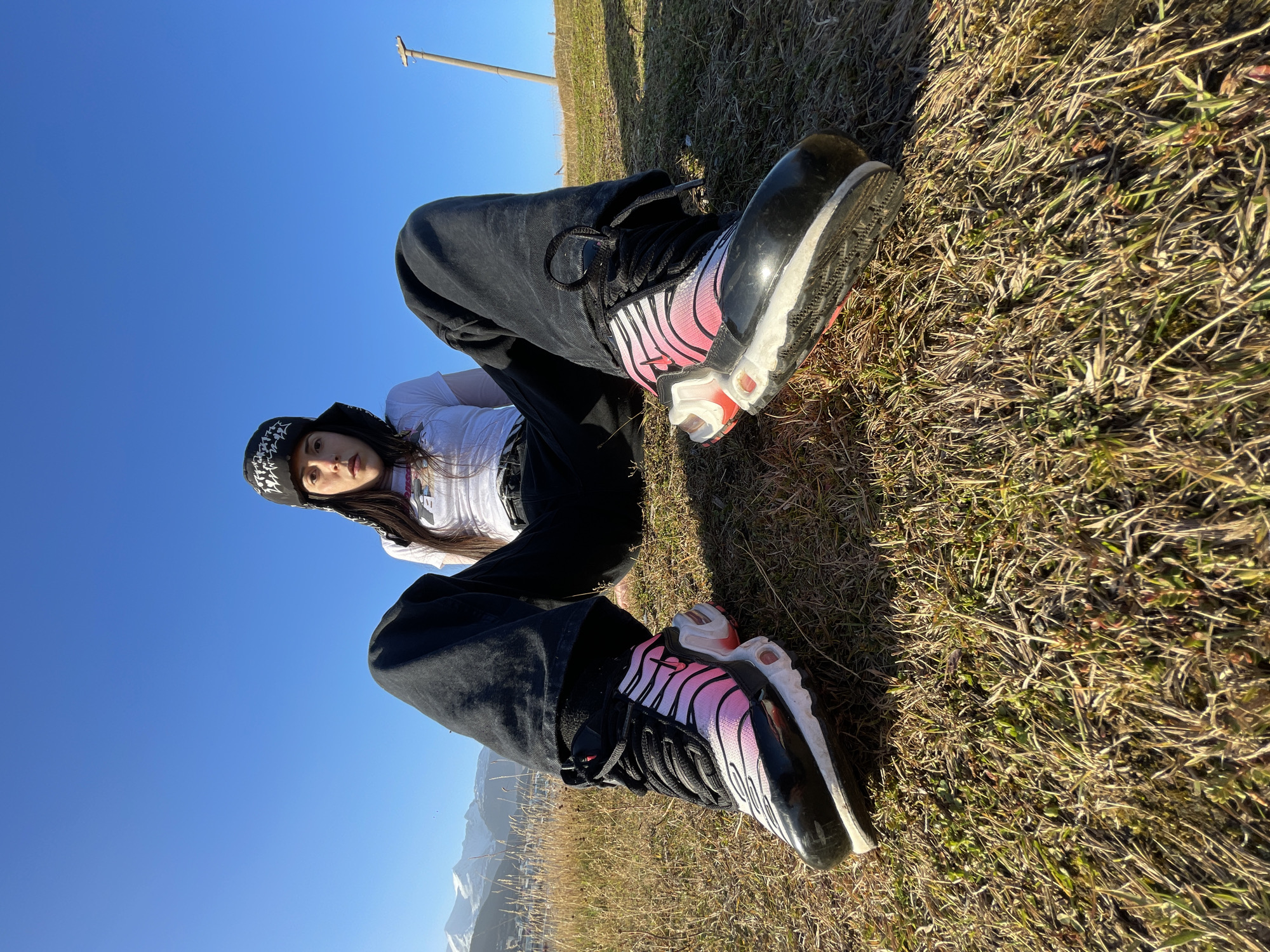 A young woman is lying on the grass, facing the camera, with her legs bent and her feet pointing towards the camera. She is wearing a white T-shirt, black pants, and a black and white patterned hat. She has long, dark hair. Her shoes are black and white sneakers with pink accents. The grass is dry and brown. In the background, there is a clear blue sky and a mountain range. There is a tall pole sticking out of the ground in the distance. The location is Ushuaia, Argentina, as suggested by the mountainous landscape typical of the region.