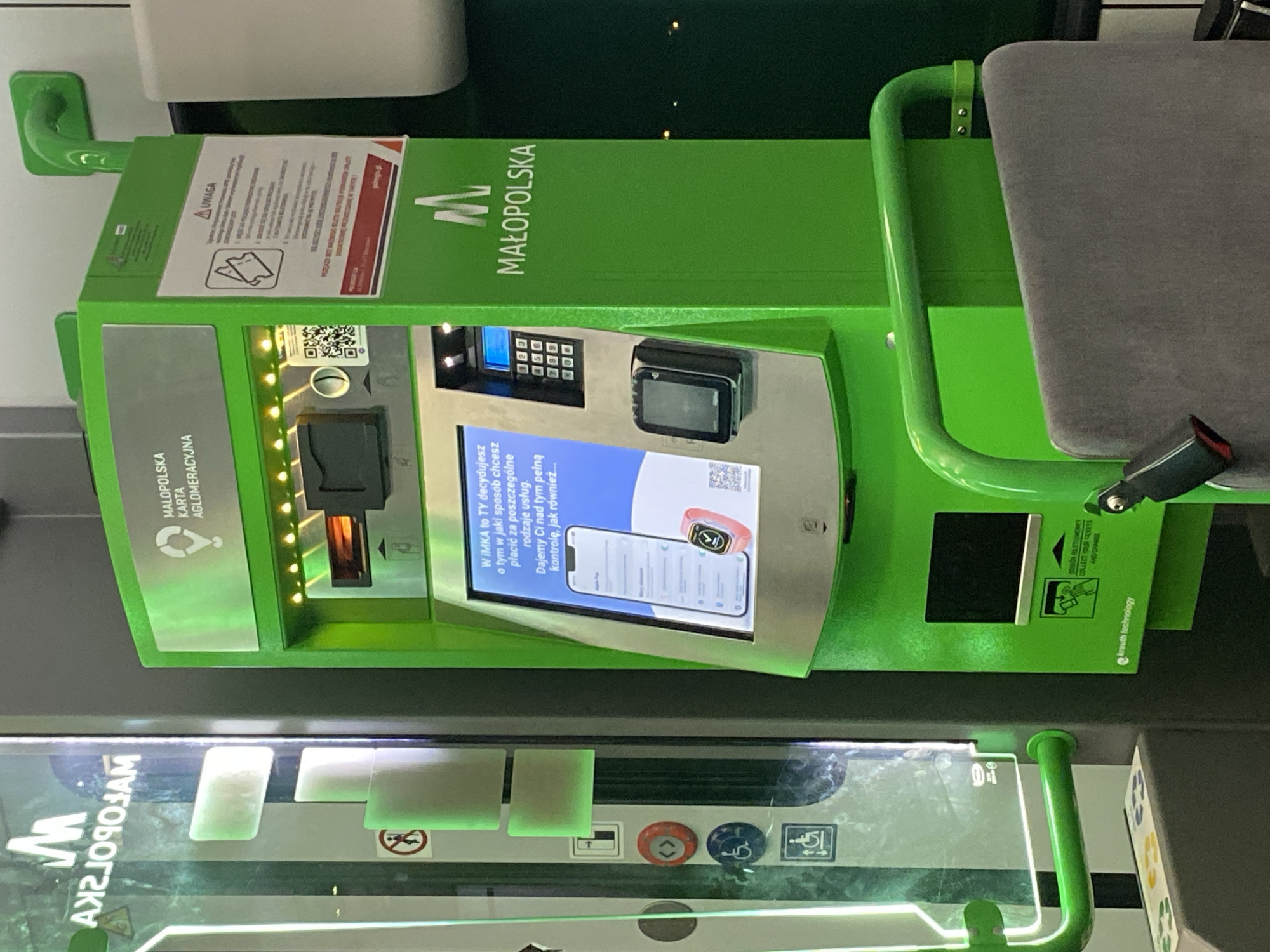 The image shows a bright green ticket vending machine manufactured by Krauth Technology, located in Łętowice, Poland. The machine has a touchscreen display showing information about payment options, including Apple Pay and an Apple Watch. There is also a keypad for entering information, a slot for inserting tickets, and a card reader. The machine is branded with the "MAŁOPOLSKA" logo, indicating it is part of the Małopolska Card public transport system.

To the left of the machine, there is a vertical display with illuminated panels, possibly a digital advertisement or information screen. This screen features the "M" logo and reversed text. There are also several buttons on the left side of the machine, including a red button with an arrow, and two blue buttons with accessibility symbols for wheelchairs.

The machine is situated in a public area, likely a transportation hub, with a metal railing in the foreground. The lighting suggests an indoor or covered outdoor environment. The machine appears to be operational, with its display and lights active.