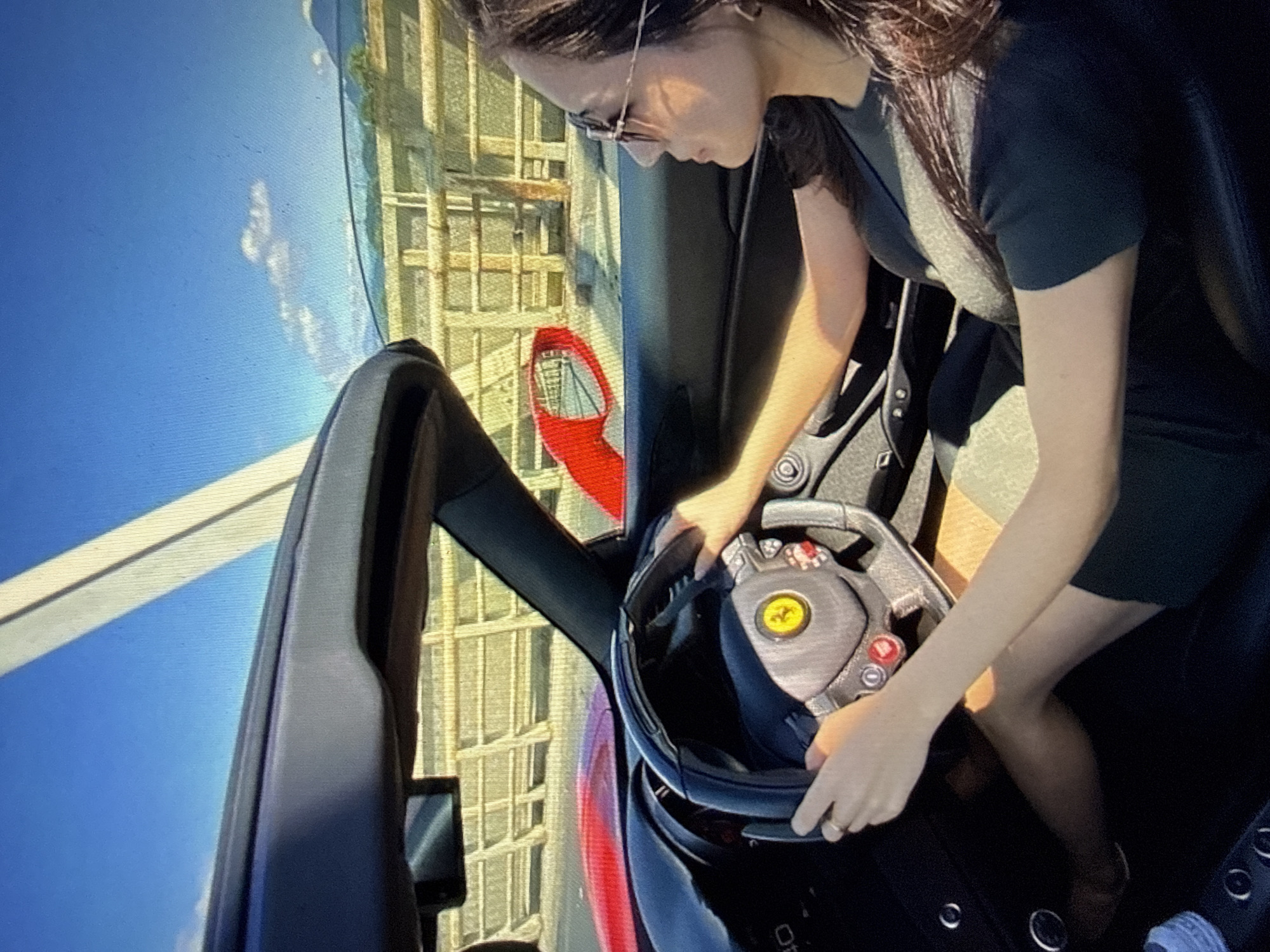 A woman is observed from the front passenger seat operating a red Ferrari convertible with the roof retracted. She has long, dark hair and wears sunglasses and a dark short-sleeved garment. Both hands are positioned on the black steering wheel, which prominently displays a yellow Ferrari logo and red control buttons; a ring is present on her left ring finger. The car's red exterior and dashboard are partially visible. A red side mirror reflects a portion of the external environment. Outside the vehicle, a substantial structure constructed from light-colored horizontal beams, indicative of a bridge or a robust barrier, extends across the view. The background consists of a clear blue sky featuring scattered white clouds and distant mountainous terrain. An unidentified white diagonal element is present in the upper sky. The depicted scene occurs in Kardzhali, Bulgaria.
