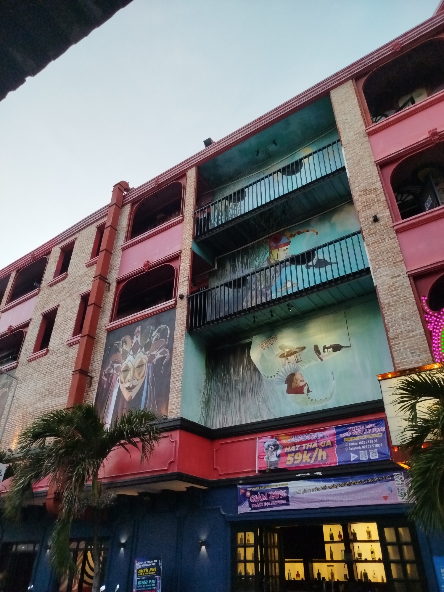 The image depicts a colorful, multi-story building in Ho Chi Minh City, Vietnam. The building's facade is a mix of red, blue, teal, and beige brick. It features arched windows and balconies with black railings. Large murals adorn the walls, including one with a Venetian mask and another with a whimsical scene of figures holding a bottle of wine. Banners are visible above the entrance, advertising karaoke ("HÁT THẢ GA") and discounts for local customers. Palm trees stand in front of the building, and lights are visible inside.