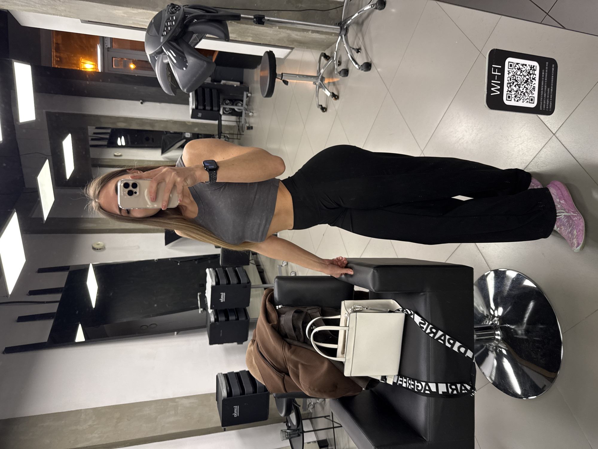 A woman stands in a hair salon, visible in a mirror. She wears a gray cropped top, black pants, and pink shoes. She holds a smartphone in her left hand, obscuring her face, and is taking a photo of herself in the mirror. Her right hand rests on a black padded chair. On the chair are a brown jacket and a white handbag with black straps. In the background, there is hair salon equipment, including hair dryers and salon chairs. A Wi-Fi sticker is visible on the floor in the upper right corner of the image. The location is inferred to be Saint Petersburg, Russia, based on the use of Cyrillic script on the black straps of the white handbag, and on the logos on the hair salon equipment behind the woman.