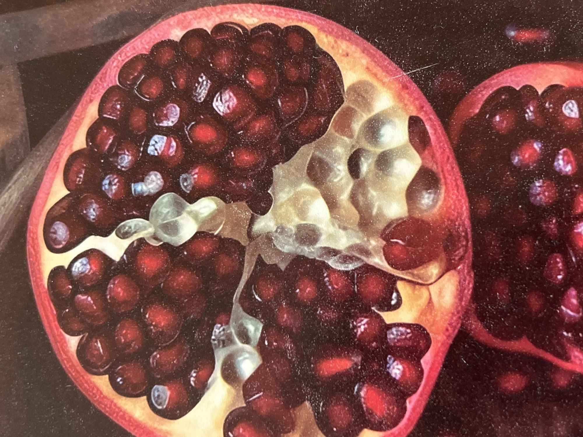 This media file features a close-up, still-life depiction of two halves of a pomegranate against a dark, textured background. The dominant subject is the fruit itself, presented in its vibrant, internal state. The left half of the pomegranate is fully visible, showcasing its jewel-like arils, a deep, rich red, clustered together. Some of the arils appear translucent, with highlights reflecting light, suggesting juiciness and freshness. The pith, a pale yellow and white membrane, is also visible, dividing the arils and adding a contrasting texture. The rind of this half is a bright pinkish-red, with a slightly rough, textured surface.
To the right, a portion of another pomegranate half is visible. It is positioned slightly behind and to the right of the first, with its rind also displaying the characteristic pinkish-red hue. The arils are less distinct in this section, but they appear to be the same deep red as in the first half.
The background is dark and appears to be a solid color, possibly a dark wood or fabric, with some subtle texture and fine specks. There are also some faint, lighter linear marks on the background, particularly on the left side, which could be reflections or part of the surface the pomegranates are resting on.
There is no indication of a specific time of day, weather, or distinct location cues beyond the implied setting of a still life composition, likely indoors in a studio or on a table. The image focuses on the visual appeal of the fruit, highlighting its textures, colors, and natural beauty. No people are visible, and no text is present in the image. The overall mood is one of natural abundance and aesthetic appreciation.