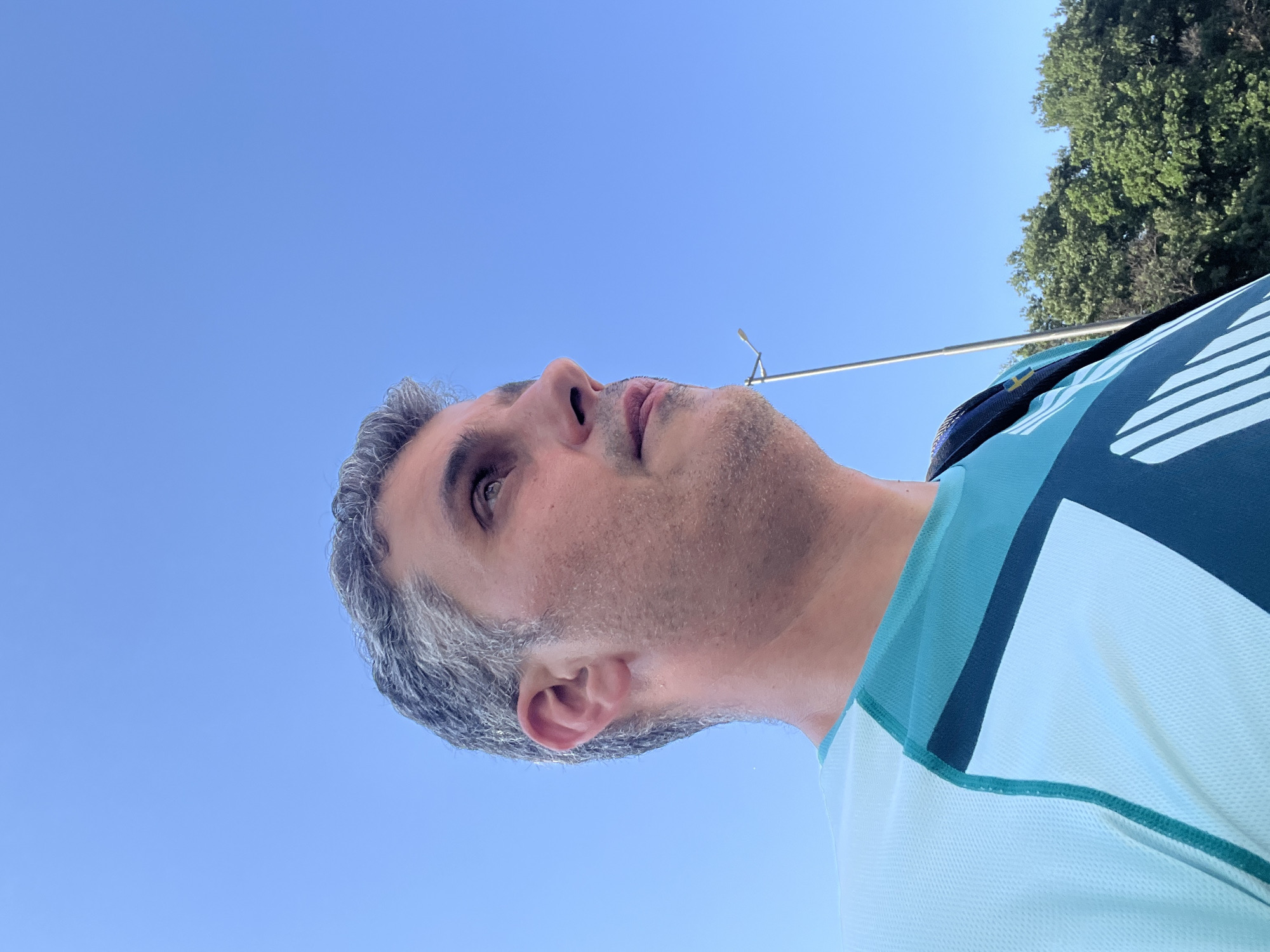 A low-angle, close-up, outdoor, medium shot captures a man looking up and to the right, against a clear, vibrant blue sky. He has short, graying hair and a light stubble on his face. He is wearing a light blue and teal athletic shirt with geometric patterns. A black strap, possibly from a backpack, is visible over his right shoulder. In the background, a tall, thin street lamp is positioned slightly off-center to the right, and a line of green trees runs along the bottom right of the frame. The image is well-lit, with the sun appearing to be behind and to the left of the photographer.