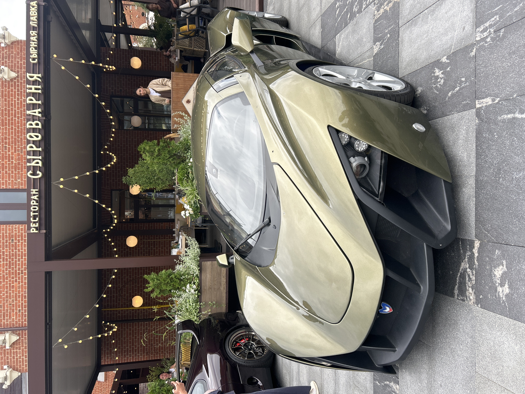 An olive green sports car is parked on a stone-tiled walkway in front of a restaurant in Moscow, Russia. The restaurant, named "СЫРОВАРНЯ" (Syrovarnya), is identifiable by its signage and the brick building it occupies. The car, with its futuristic design, occupies the foreground of the image. Behind the car, to the left, a dark colored sports car is partially visible, with a large spoiler. The restaurant features an outdoor seating area with tables and chairs, and is decorated with string lights. Several people are visible in the background, including a woman standing behind a counter inside the restaurant and other patrons seated at tables. The ground is paved with large, irregularly shaped stone tiles.
