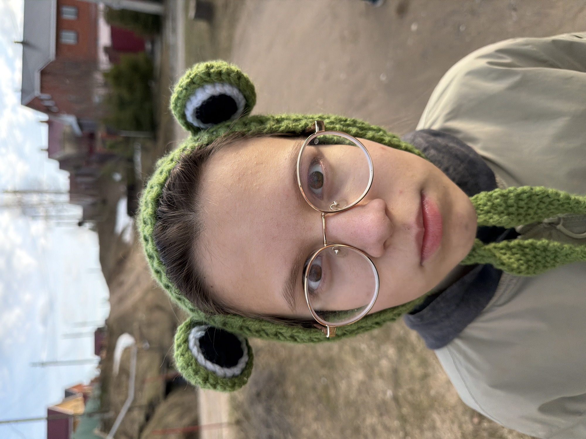 A close-up, portrait-style photograph captures a young person from the chest up, positioned against a blurred outdoor background. The subject, likely a teenager or young adult, faces the camera directly with a neutral expression. They wear distinctive round, gold-rimmed glasses that sit on their nose, reflecting light. Their eyes are brown.
A prominent feature is the green crocheted hat, styled to resemble a frog, with large, round eyes protruding from the top, featuring black centers and white rings. The hat's textured, knitted material is clearly visible. The subject also wears a light olive-green or khaki-colored jacket, with a dark grey inner layer or collar visible around the neck.
The background suggests an outdoor, possibly residential or rural setting in Chaikovsky, Russia. Blurred houses with sloped roofs, some reddish-brown and others lighter, are visible. Bare trees or sparse vegetation are also present, hinting at cooler weather, possibly late winter or early spring. The sky appears bright but overcast, providing soft, diffuse lighting. No text is visible in the image.