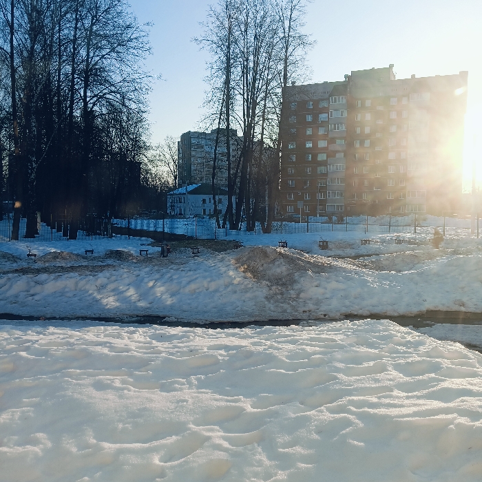 The scene depicts an urban winter or early spring landscape in Chaikovsky, Russia, under a clear, bright sky. The foreground is occupied by an undulating expanse of fresh, undisturbed white snow. In the midground, a snow-covered embankment or slope presents patches of exposed dark earth and several small, rectangular wooden structures, possibly benches or equipment bases, sparsely distributed across it. A light-colored, low fence runs horizontally behind this embankment. The background features a dense cluster of tall, bare deciduous trees. Through the trees on the left, a white two-story building with a dark roof and a taller, multi-story grey apartment building are visible. On the right, a prominent multi-story brick apartment building with numerous windows and balconies stands against the bright sky. Intense, low-angle sunlight emanates from the right side of the image, causing significant glare and partially obscuring the right side of the brick building. No individuals or discernible activities are present within the frame.
