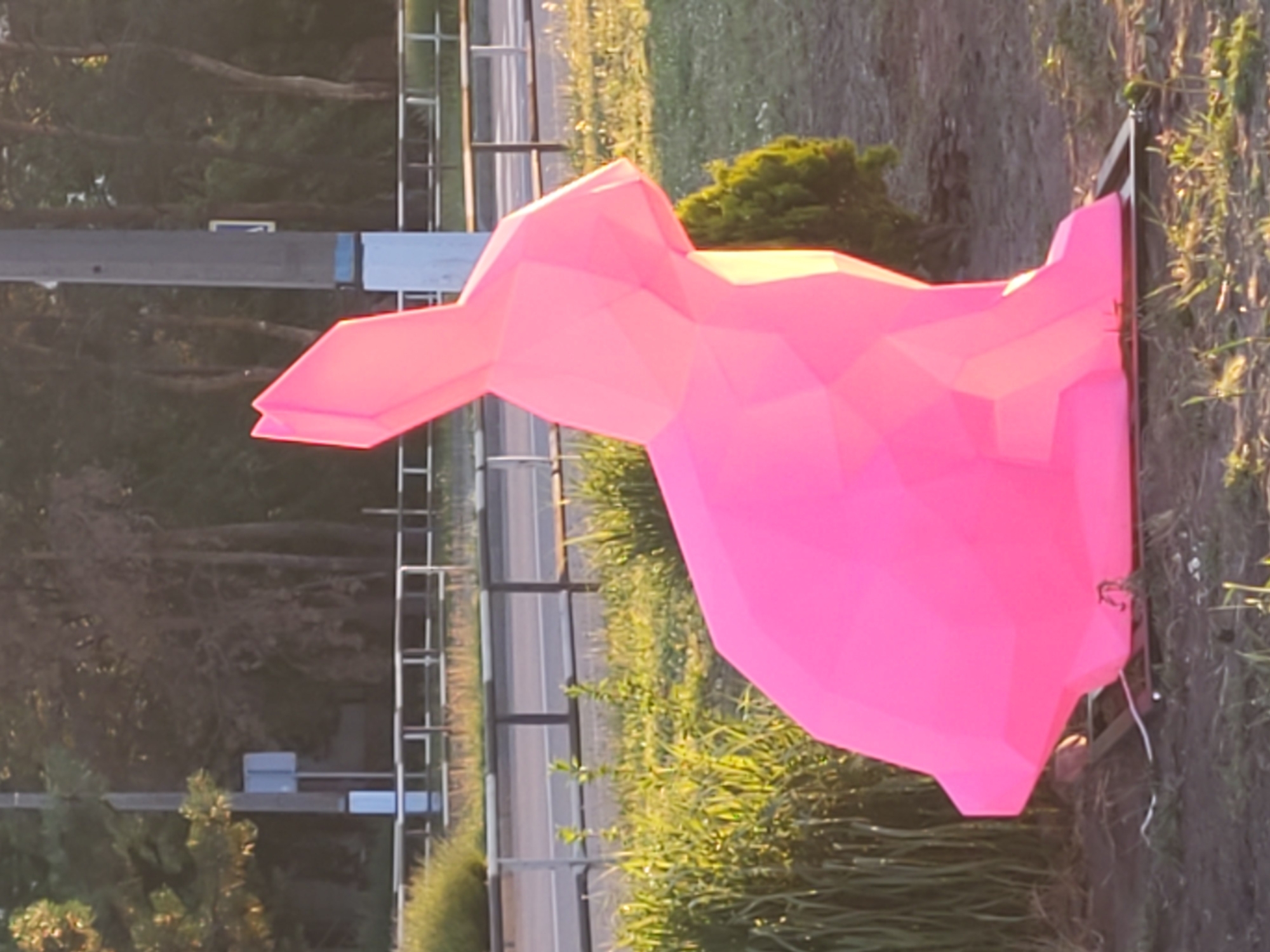 A neon pink, polygonal sculpture of a rabbit is positioned on a metal base in the foreground, partially obscuring grassy ground and some dirt. The sculpture's geometric design is evident, with sharp angles and flat planes. Behind the sculpture, a metal railing with white and black bars runs horizontally, separating the area from a road. A paved road with white lines marking lanes is visible beyond the railing. A grey utility pole stands to the right of the rabbit sculpture, with a blue and white sign attached to it. Trees with green foliage and some bare branches form a backdrop, with dappled sunlight suggesting an outdoor environment. The overall scene depicts a public area in Horishni Plavni, Ukraine, featuring a distinctive art installation.