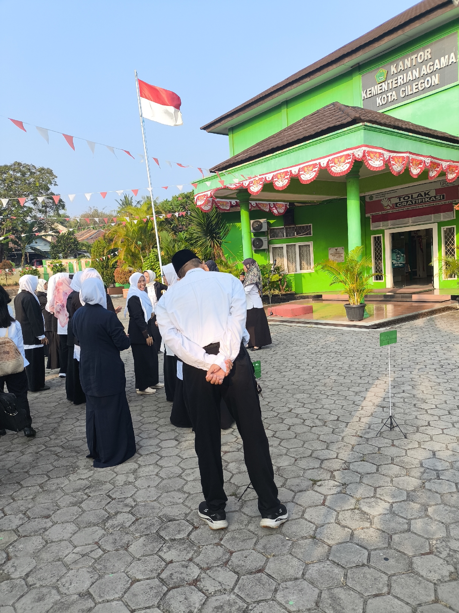 The image depicts an outdoor scene in front of a green building with the sign "KANTOR KEMENTERIAN AGAMA KOTA CILEGON" which translates to "Office of the Ministry of Religious Affairs, Cilegon City". The building has a dark tiled roof and is decorated with red and white pennant banners along the eaves. An Indonesian flag, red and white, is prominently displayed on a flagpole to the left of the building. A group of people, mostly wearing dark uniforms and headscarves, are gathered in front of the building on a hexagonal paved courtyard. Some are facing the building, while others are turned away, with one individual in the foreground facing away from the camera, hands clasped behind their back. A potted plant sits near the entrance of the building. The sky is clear and blue, and the sun casts shadows on the pavement. The environment appears to be a public or governmental facility, likely during a morning gathering or ceremony given the flag and banners.