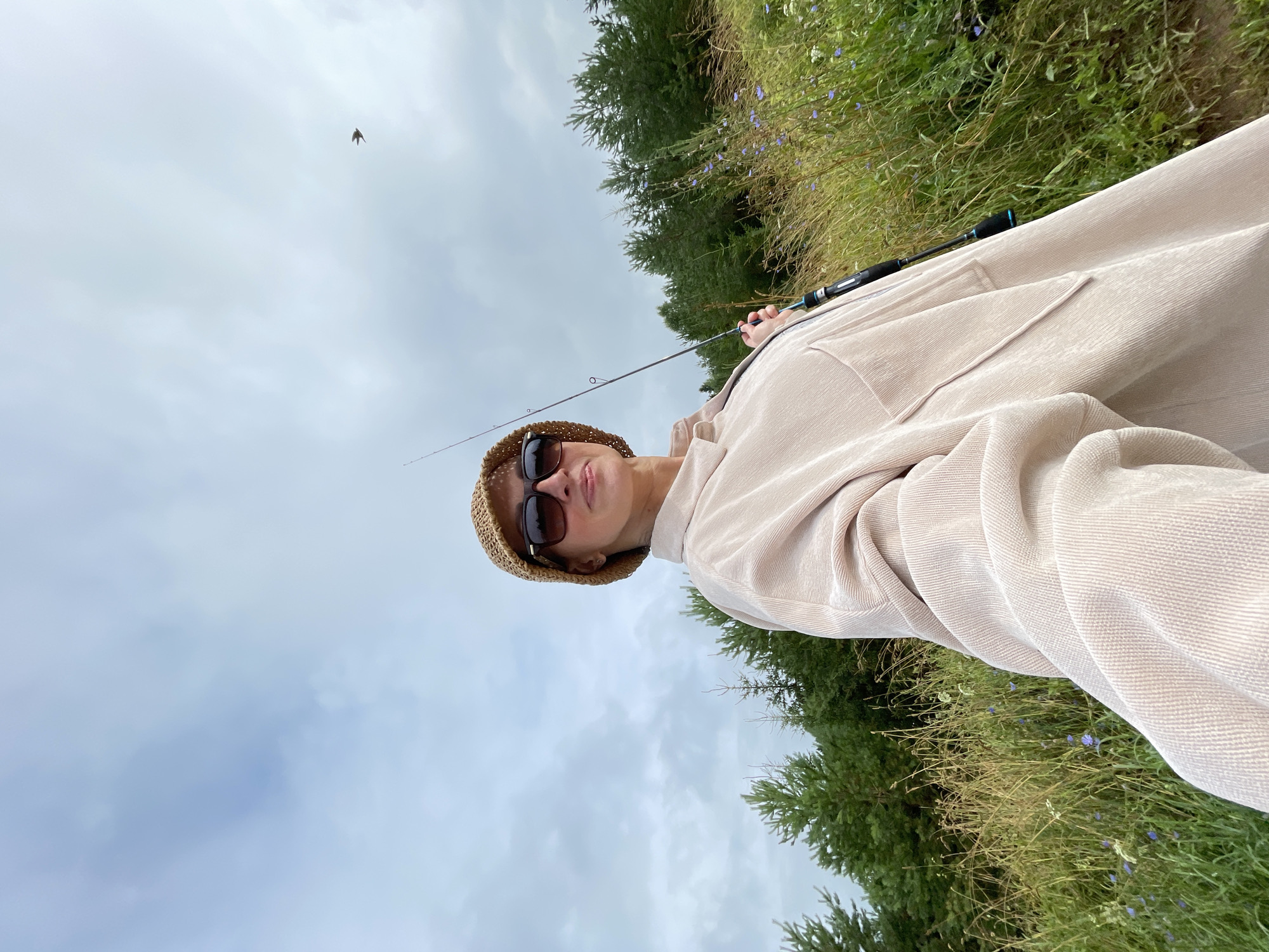 A woman wearing a light-colored oversized shirt, a straw hat, and sunglasses is holding a fishing rod. She is outdoors in a grassy area with trees in the background. The sky is cloudy. A small bird is visible in flight in the upper right corner of the image. The scene appears to be a natural landscape, possibly near a body of water, in or around the city of Bereznik, Russia.