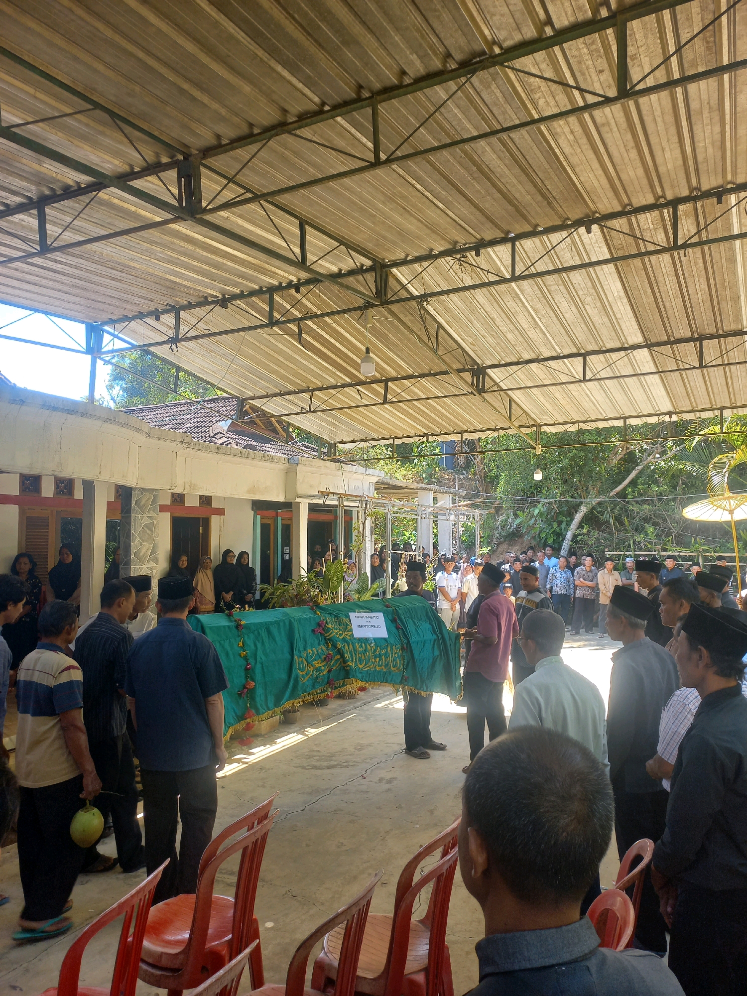 The image depicts a funeral procession under a covered outdoor area in Sukoharjo, Indonesia. A coffin, draped in a green cloth with gold lettering that reads "HADJI SABITO BIN MARTOREJO," is the central focus. A crowd of people, mostly men wearing traditional Indonesian attire like peci caps, are gathered around the coffin and further back. The environment appears to be an open space with a corrugated metal roof supported by a metal structure, suggesting a community hall or outdoor gathering space. A building with windows and doors is visible in the background, and lush green trees and foliage surround the area, indicating a rural or semi-rural setting. The lighting suggests it is daytime, with sunlight casting shadows on the ground. Some red plastic chairs are visible in the foreground, suggesting a seating arrangement for mourners. One man in the foreground is holding a green coconut. The overall atmosphere is somber and respectful, typical of a funeral gathering.