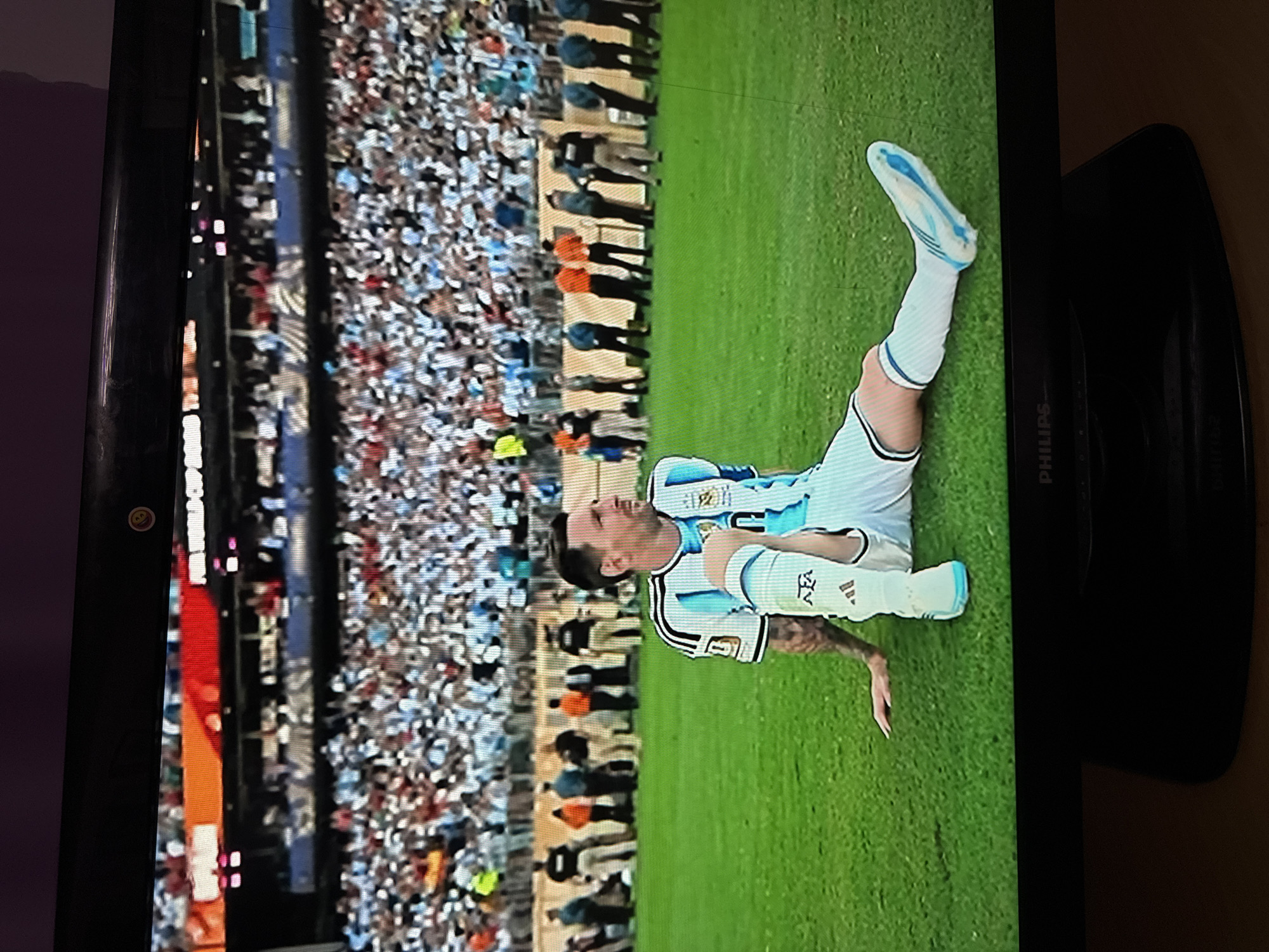 The image shows a television screen displaying a soccer player, Lionel Messi, sitting on a grassy field. He is wearing a white and light blue Argentina national team jersey and shorts, with white socks and cleats. The field is green, and in the background, a large crowd of spectators is visible, seated in stadium stands. Above the crowd, a banner with "WORLD CUP" is partially visible. The scene is illuminated by stadium lights. The television itself is black, with a "PHILIPS" logo visible on the bottom bezel. The environment outside the screen suggests it is being viewed in a room with wooden furniture. The image does not provide any information about the city of Ufa, Russia.
