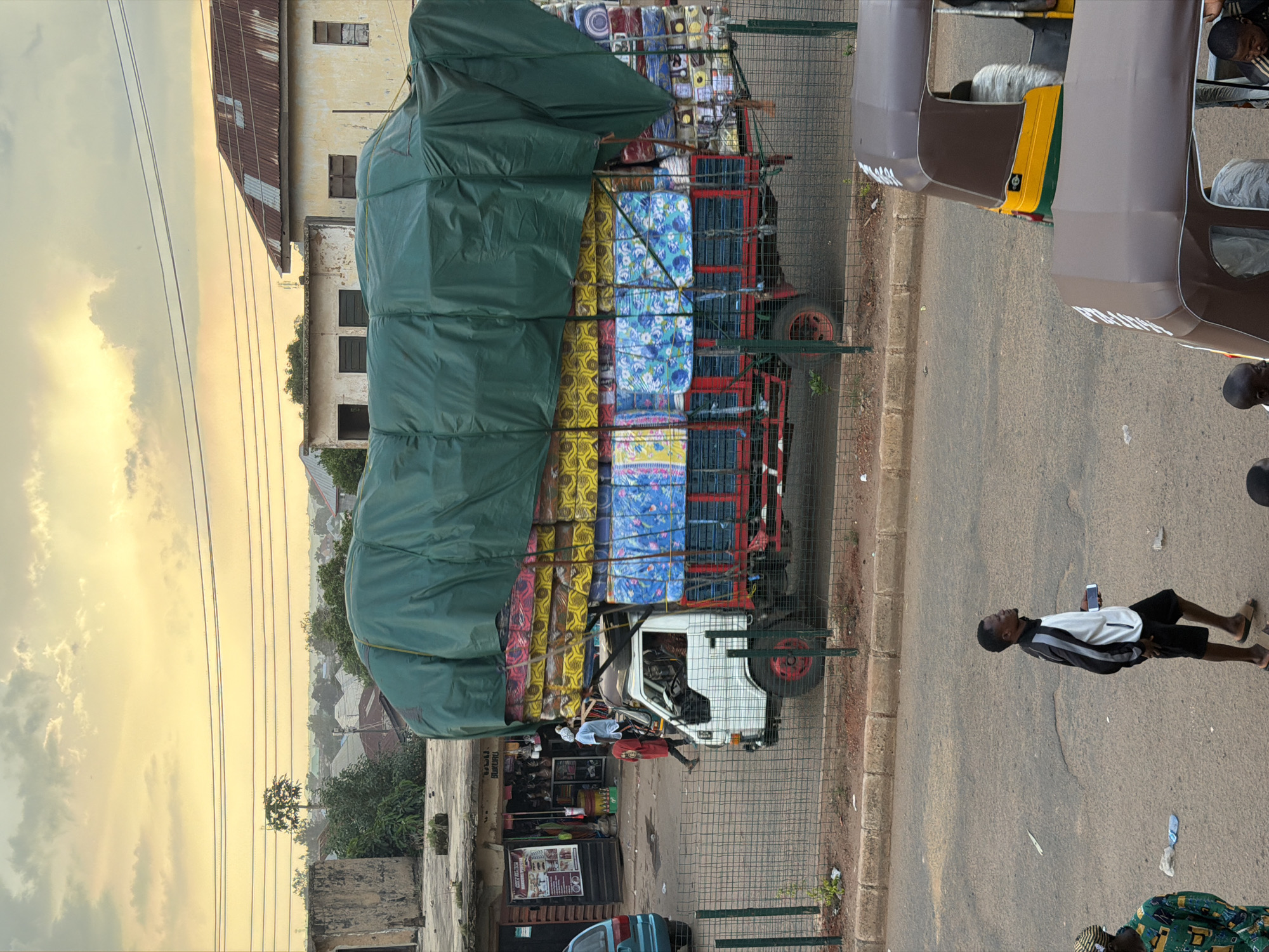 In Bukuru, Nigeria, a flatbed truck is heavily laden with mattresses, many of which are wrapped in plastic and colorful patterned fabrics. The cargo is secured with a large, dark green tarp. A white truck cab is visible, with a person in a red shirt walking near the driver's side. In the background, buildings with corrugated metal roofs and some with concrete structures line the street. The sky above is a pale yellow and orange, suggesting either sunrise or sunset. In the foreground, two people are visible walking on the road. One is wearing a black and white tracksuit and black shorts, holding a phone, and the other is wearing a green and yellow patterned shirt. Two auto-rickshaws are parked on the right side of the image. A wire fence separates the road from where the truck is parked.