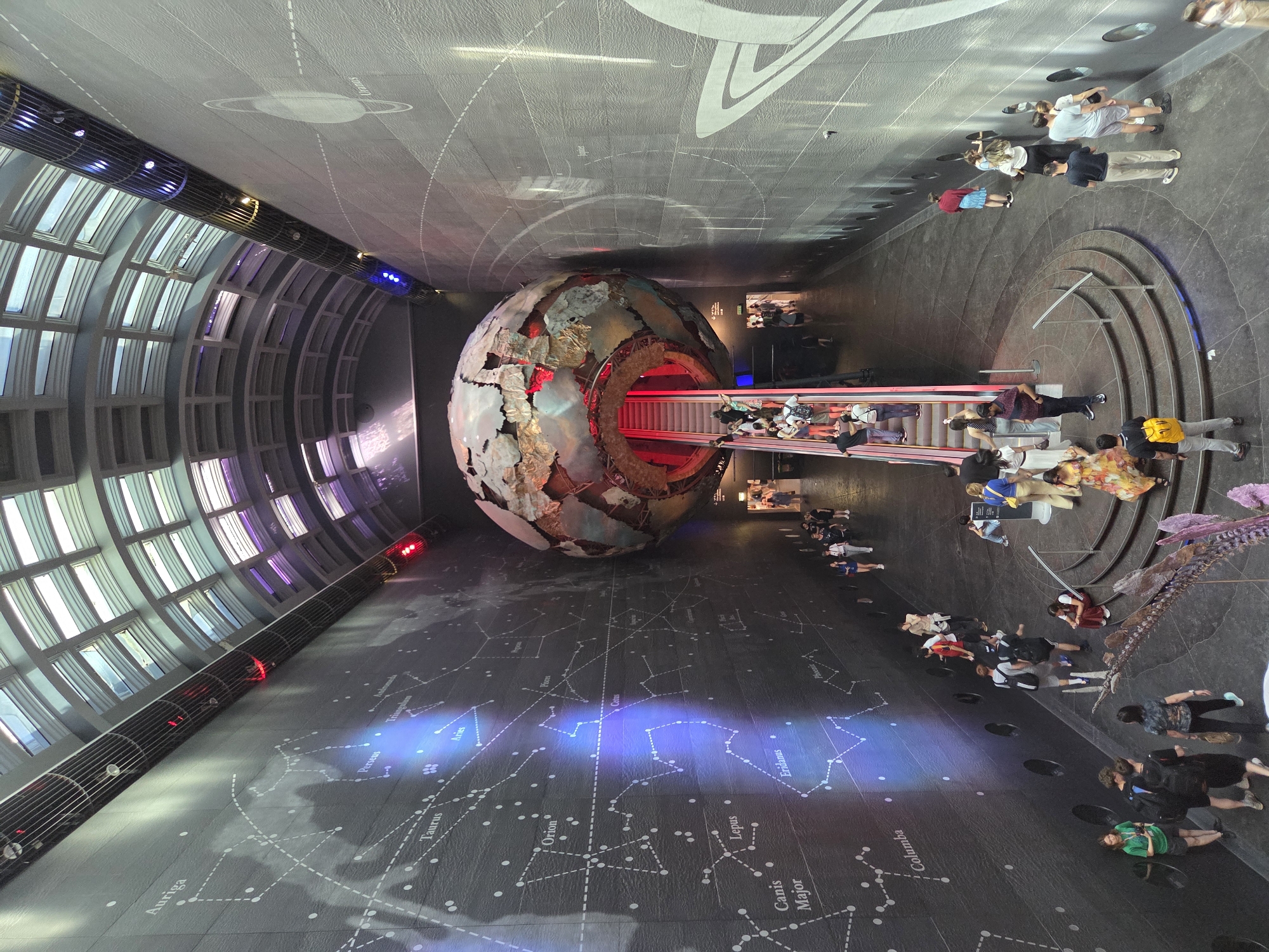 The image depicts an interior view of the Hintze Hall in the Natural History Museum in London, United Kingdom. The hall features a large metal globe structure, a cross-section of Earth, positioned prominently in the center. A red-lit escalator cuts through the globe. A throng of people are seen both around and on the escalator.
The hall is characterized by a long, arched window structure on one side, composed of numerous individual panes. The floor displays a constellation map rendered in white lines and dots over a dark surface. The ceiling is painted with planetary diagrams. The individuals in the hall are a mix of adults and children, dressed in casual clothing, moving around and observing the exhibits.