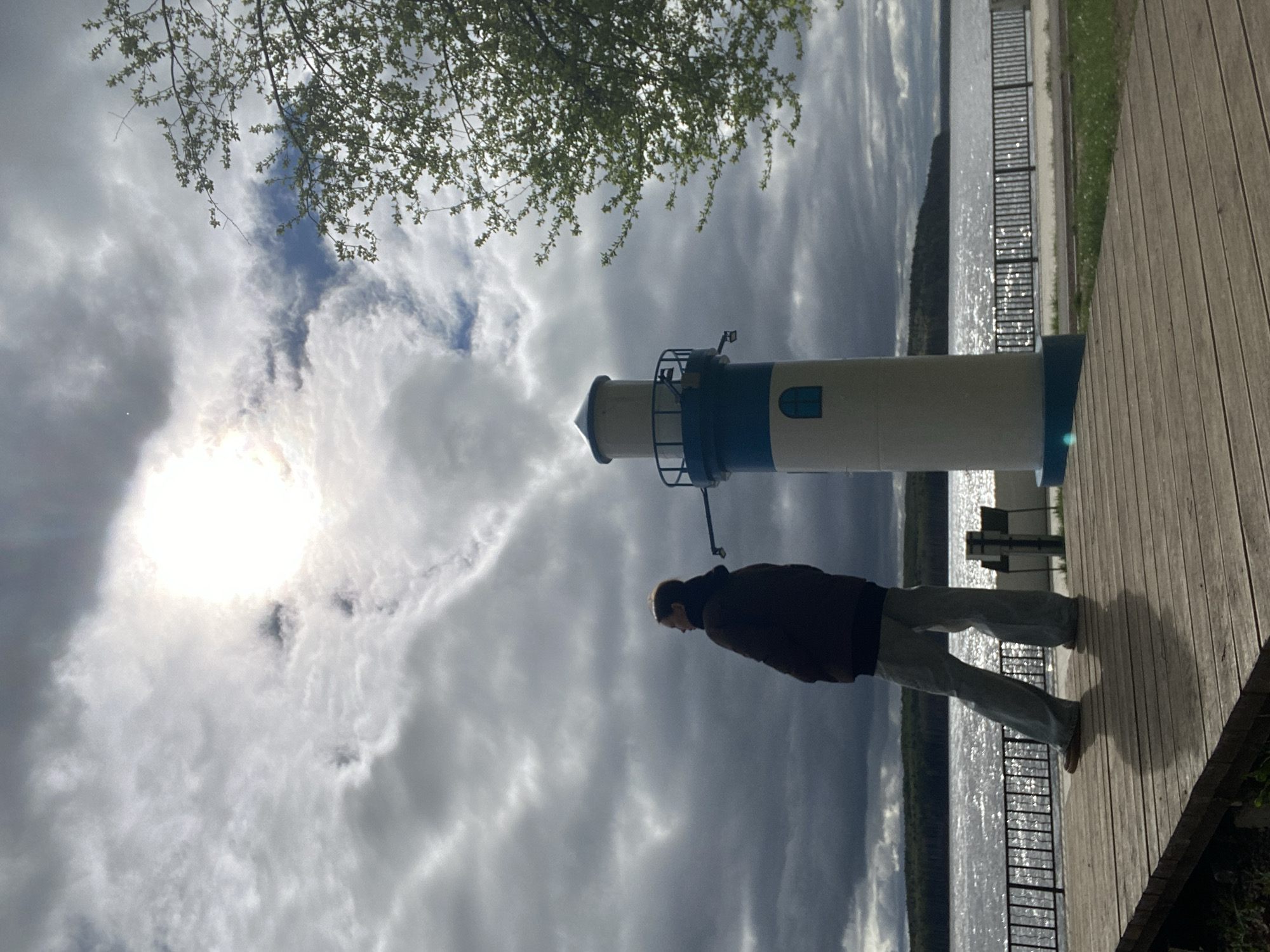 A long shot shows a man standing on a wooden deck next to a replica lighthouse. The lighthouse has a blue top and base, with a white body and small blue windows. A metal railing encircles the top. The man wears a dark jacket and light-colored pants. A body of water lies behind the deck, reflecting sunlight. Beyond the water is a forested area. Overhead, a sky filled with grey and white clouds is partly obscured by branches with green leaves. The location is Chaikovsky, Russia.