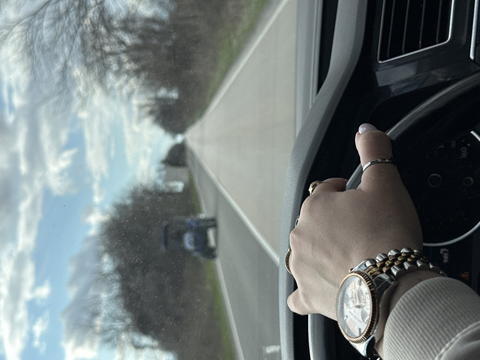 The image depicts a first-person perspective from the driver's seat of a vehicle traveling on an asphalt road in Kvareli, Georgia. The driver's right hand, adorned with a two-tone wristwatch (silver and rose-gold with a white dial and chronograph subdials), a gold ring on the ring finger, a second gold ring on the middle finger, and a simple silver band on the thumb, grips the top-left portion of the black steering wheel. The driver's arm is covered by a light-colored sleeve. Through the windshield, a two-lane road with a broken white center line and a solid white shoulder line recedes into the distance. Ahead, a blue, open-cab vehicle, possibly a small tractor or utility cart, is visible in the same lane, moving away from the viewer. Bare deciduous trees line both sides of the road. A small, light-colored structure is partially visible on the left side of the road beyond the blue vehicle. The sky above is partly cloudy with patches of blue. The dashboard, with a visible air vent on the right, is dark-colored.