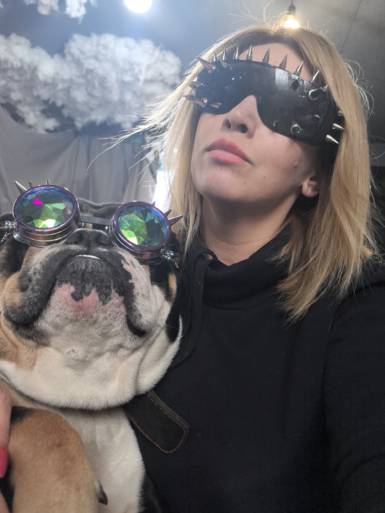 An indoor, close-up shot features a woman and a bulldog, likely captured in Noviy, Russia. The woman, with blonde hair, wears a black hooded top and a striking black eye mask adorned with numerous metallic spikes. Her lips are painted with pink lipstick, and she has a calm, confident expression, looking slightly upwards and to the right. She is holding a bulldog, which also wears unique eyewear: steampunk-style goggles with iridescent, multifaceted lenses and small spikes on the frame. The bulldog's face is mostly white and brown, and it appears to be held snugly by the woman. In the background, there are cloud-like white decorations, possibly on a ceiling or wall, and a bright light source creating a lens flare effect, suggesting an artificially lit or creative environment. There is no visible text in the image.