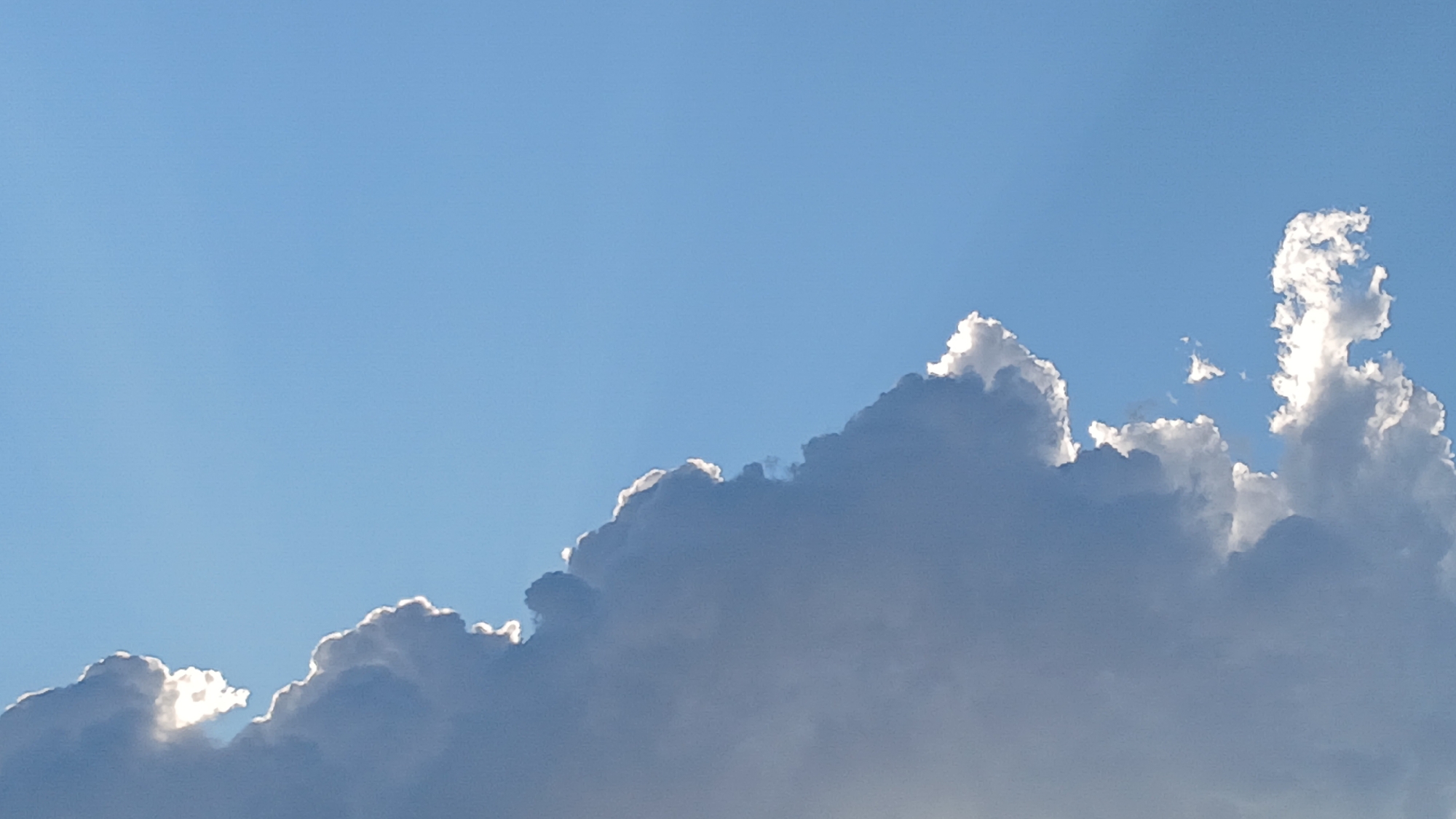 A wide shot captures a bright blue sky with a line of dramatic, sunlit clouds stretching across the frame from the bottom right to the top left. The clouds are a mix of dark gray and white, with the edges brightly illuminated by the sun, creating a luminous halo effect. The sky is clear and cloudless to the left, creating a stark contrast with the textured clouds on the right. No specific geographical landmarks of Astana, Kazakhstan, are visible in this image.