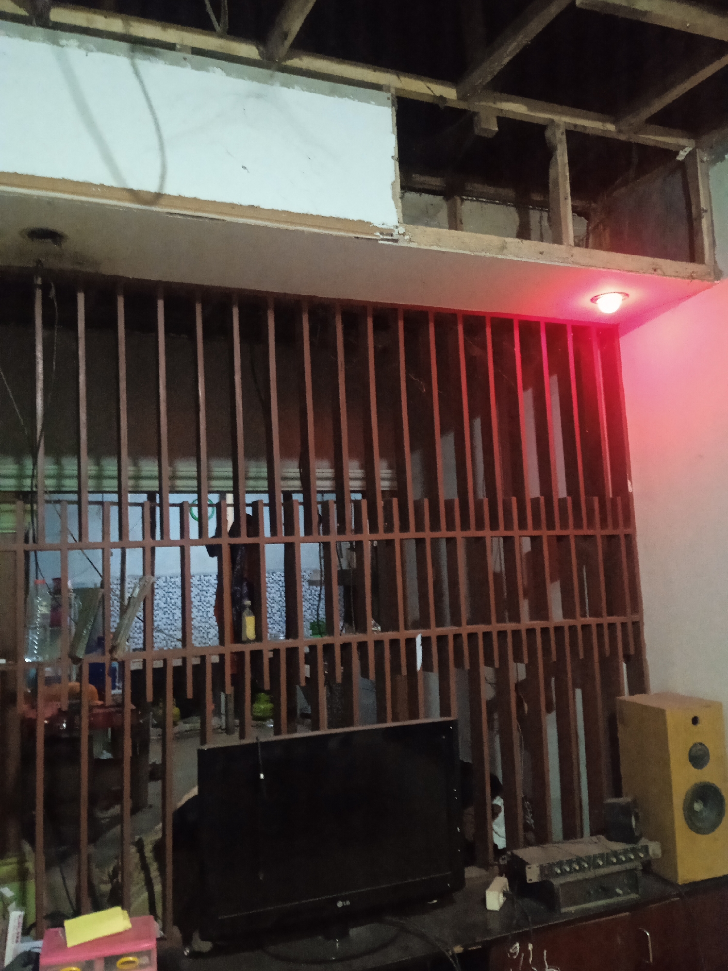 The image depicts an interior scene in Jatisrono, Indonesia, characterized by a rustic and unfinished aesthetic. In the foreground, a black flat-screen television sits on a dark wooden surface, likely a cabinet or desk. To the right of the television is a light brown speaker cabinet. In front of the speaker and television, a metallic device with numerous knobs and buttons is visible, possibly audio equipment. Further to the right, a smaller black speaker rests on the same surface.
Dominating the midground is a wooden grille or partition made of vertical slats, suggesting a separation of spaces. Through the grille, a tiled wall with a blue and white geometric pattern is partially visible, along with various items, including bottles and what appears to be clothing or fabric draped over a person's shoulder. Behind the grille, the ceiling is unfinished, exposing wooden beams and electrical wiring. A white ceiling panel with a rough edge is also present.
A red light fixture is mounted on the underside of a wooden beam above the grille, casting a red glow on the scene. The overall environment suggests a living space or workshop undergoing renovation or construction, with a focus on practical functionality rather than decorative finish. The lighting is dim, with the red fixture providing an accent illumination. The image is taken from a slightly elevated angle, looking down at the television and speakers.
