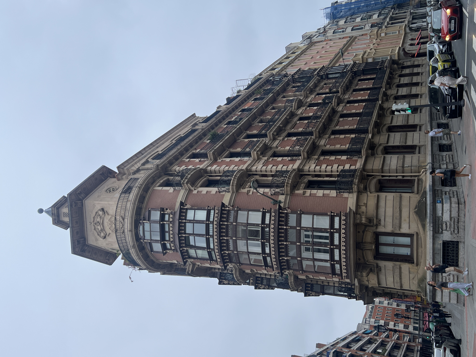A low-angle, full-shot captures a corner building in Bilbao, Spain, with an overcast sky. The building is made of reddish-brown brick and has ornate architectural details, including bay windows with decorative ironwork, balconies with black metal railings, and a tiered, octagonal tower at the corner. Several people are walking on the sidewalk in front of the building, and cars are parked and driving on the street. The building to the right is pink and has similar architectural features. The scene appears to be a typical urban streetscape in Bilbao, likely in the city center.