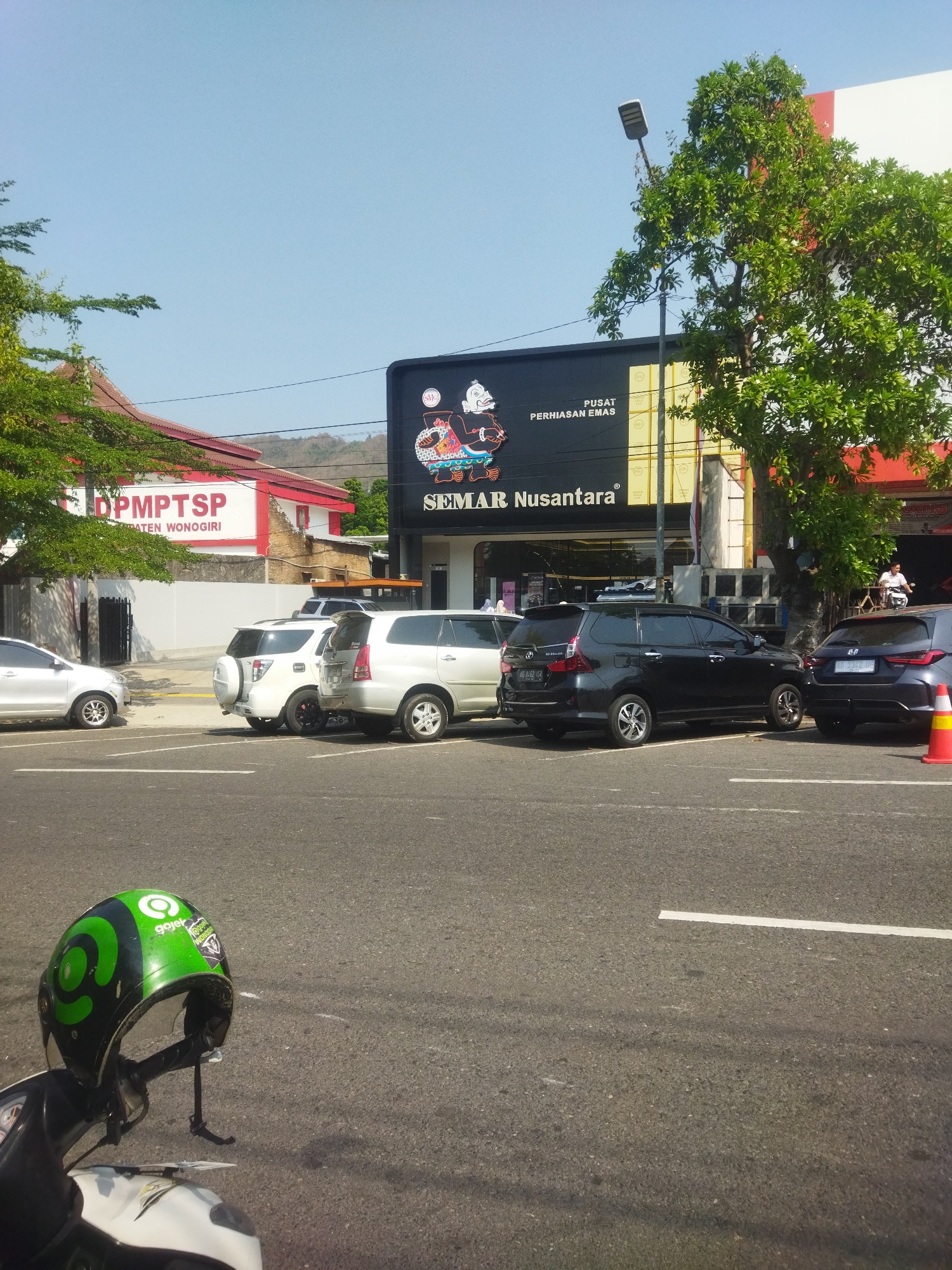 In the city of Wonogiri, Indonesia, a street scene unfolds under a clear blue sky. In the foreground, a green and black Gojek helmet rests on the handlebars of a partially visible white scooter. The street is paved with asphalt and marked with white lines.
Dominating the mid-ground are several parked cars. A silver sedan is positioned on the left, followed by two white SUVs. Further to the right, a black minivan is parked, and beside it, a dark-colored SUV. A bright orange traffic cone is visible on the right side of the frame.
In the background, a building with a black facade displays a large sign that reads "PUSAT PERHIASAN EMAS SEMAR Nusantara". The building is flanked by a tree with lush green foliage on the right. To the left of the "Semar Nusantara" building, another structure with a red roof and white walls is visible, featuring red lettering that appears to say "DPMPTSP KABUPATEN WONOGIRI".
A street lamp stands tall next to the "Semar Nusantara" building. In the far distance, a gently sloping hill is visible under the bright sunlight. A person on a motorcycle can be seen in the background, near the entrance of the "Semar Nusantara" building.
