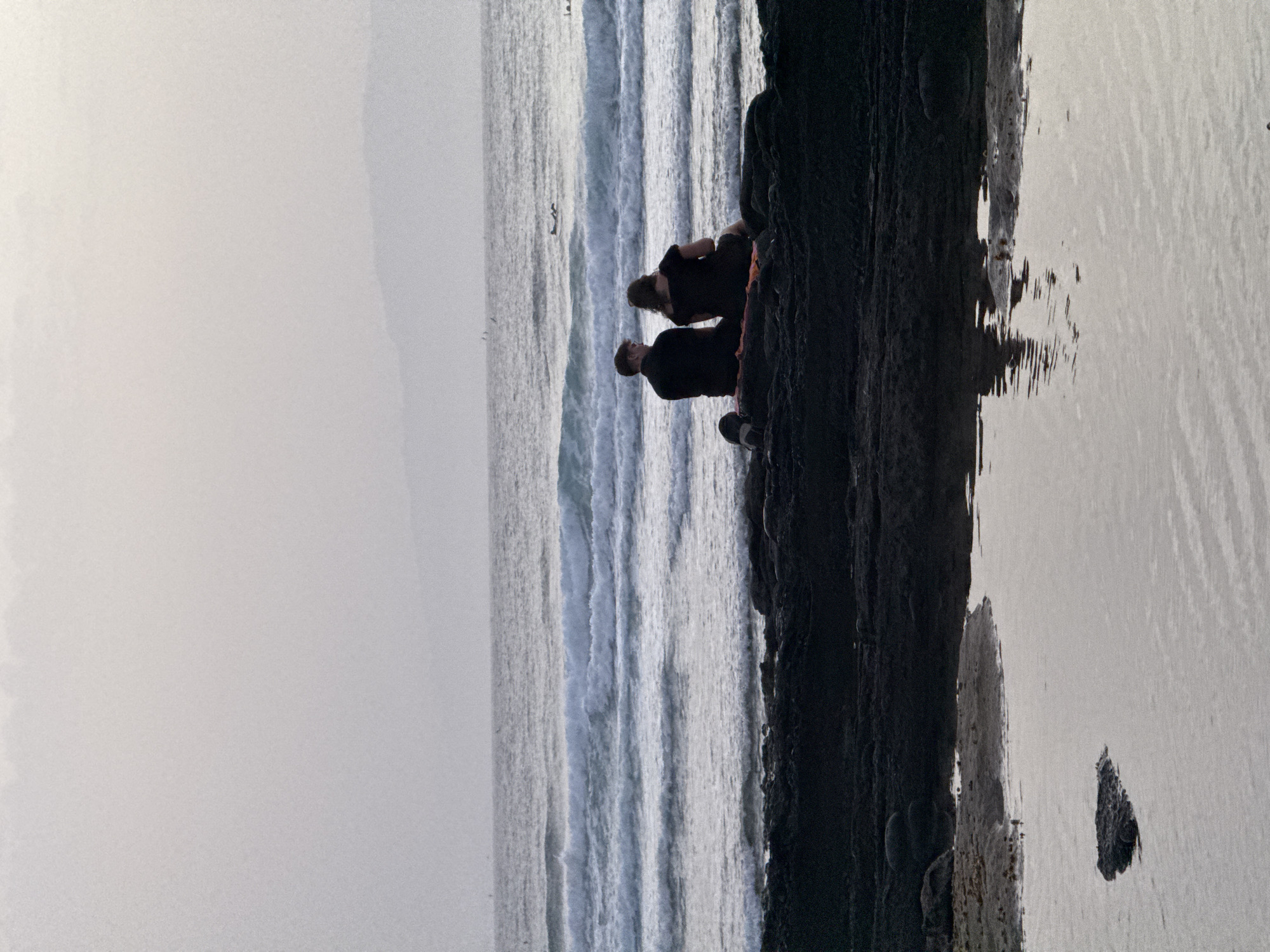 An image shows two people, a man and a woman, sitting on dark volcanic rocks facing the ocean. The man wears a dark shirt and trousers, and the woman wears a dark dress. They are sitting side by side on a reddish blanket. The ocean is calm with small waves, and a distant coastline is visible on the horizon. The sky is overcast and light gray. This scene appears to be at a beach in Arona, Spain, characterized by its black volcanic rock formations. In the foreground, a shallow pool of water reflects the sky and rocks. Several surfers are visible in the distance on the ocean waves.
