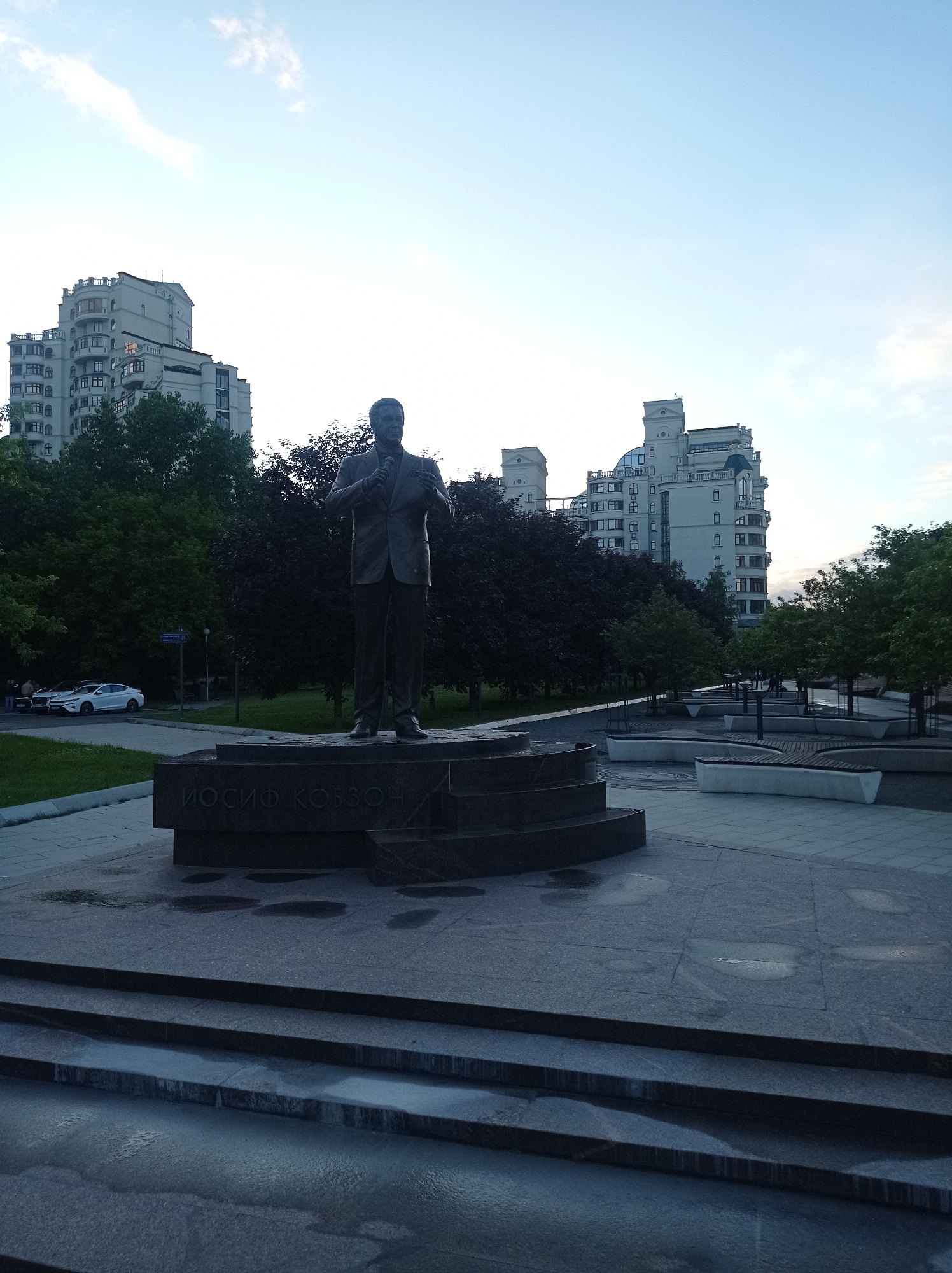 In a public plaza in Moscow, Russia, a bronze statue of Iosif Kobzon stands on a granite pedestal. The statue depicts Kobzon in a suit, holding a microphone. The plaza is paved with granite tiles and features tiered seating areas and decorative fountains. In the background, modern apartment buildings and trees are visible. The sky is clear with a few wispy clouds. The scene appears to be captured in the late afternoon or early evening.