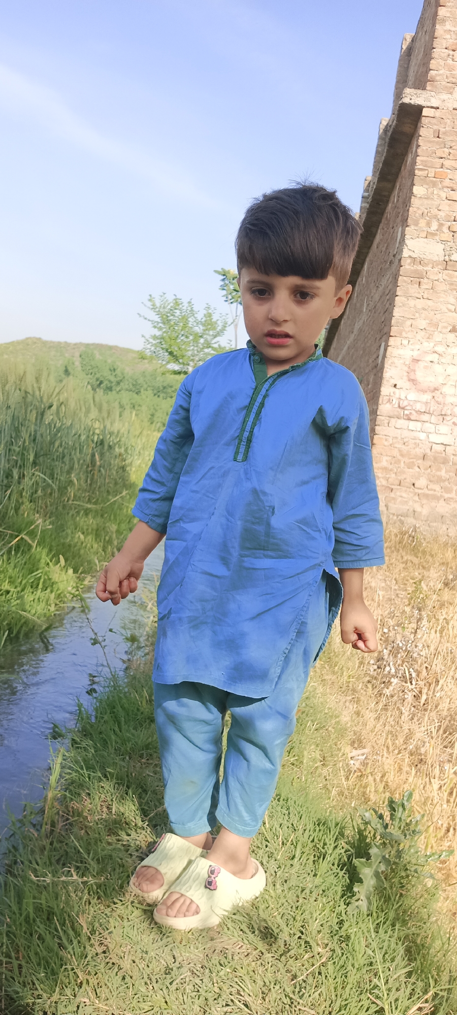 A young boy stands in a grassy field, wearing a blue kurta shalwar with green trim at the neck. He is positioned near a small stream, with a field of wheat or similar grain visible in the background. A brick structure is partially visible behind him to the right. He is wearing light-colored sandals. The location appears to be a rural area.