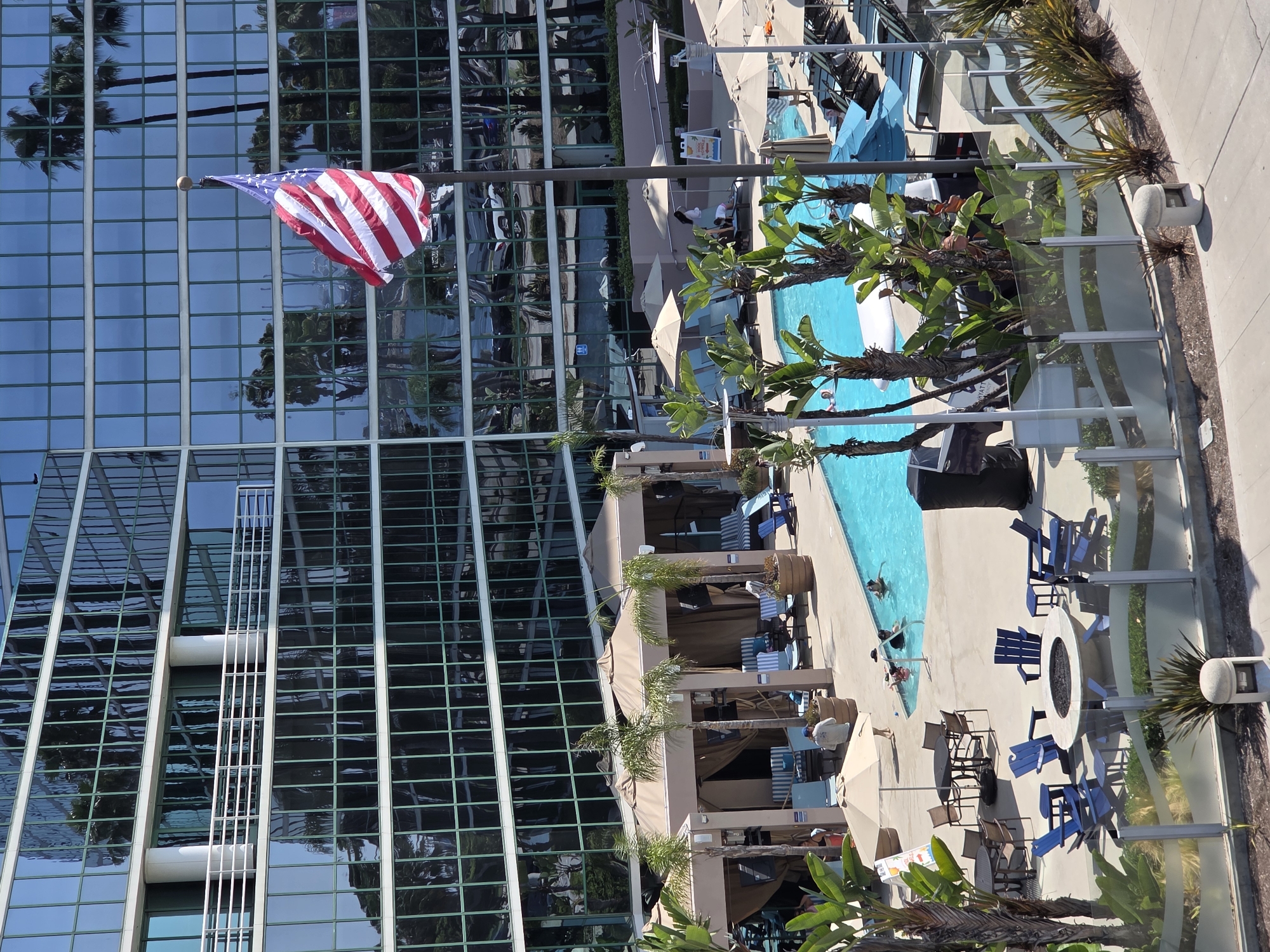 This is an outdoor, high-angle photograph of a hotel courtyard in Long Beach, California. In the background, a modern, multi-story building with a glass facade reflects palm trees and a clear sky. In the middle ground, an American flag waves from a flagpole in front of the building. To the left, a swimming pool is visible, with several people swimming and relaxing in the water. Around the pool are lounge chairs, umbrellas, and cabanas. There is also a fire pit area with blue Adirondack chairs and a round stone table. In the foreground, a paved walkway and a glass fence separate the courtyard from the viewer. The scene is bathed in sunlight, indicating it is daytime.