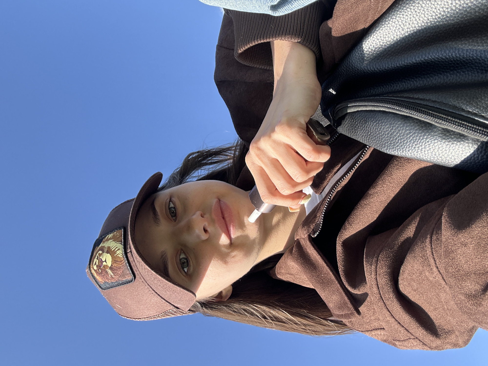 A woman wearing a brown baseball cap with a lion patch, a brown jacket, and a black bag is applying lip gloss. The sky is clear and blue. There are no identifiable features to indicate this photo was taken in Izhevsk, Russia.