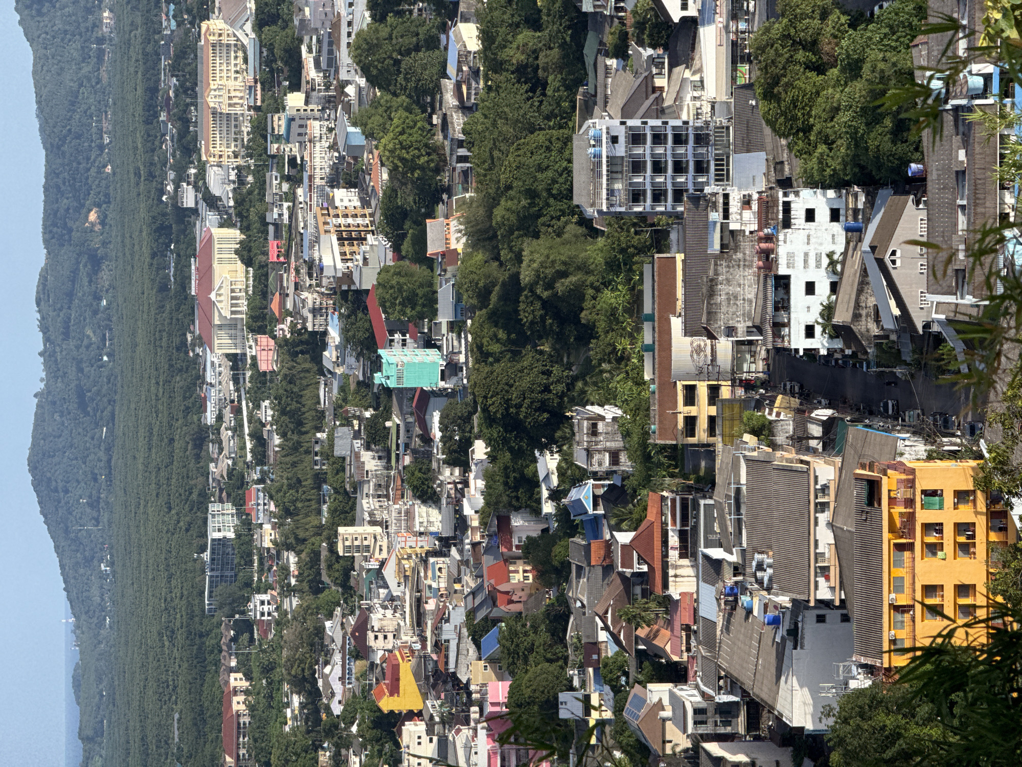An aerial, high-angle, long shot shows the densely populated city of Ratsada, Thailand. The city is nestled between a large, lush green mountain range and the ocean, visible as a thin blue line in the distance. The foreground is dominated by the rooftops of various buildings, including residential homes and commercial structures. Buildings vary in height and architectural style, with many featuring multi-colored facades, predominantly yellow, orange, and white. Some buildings have visible air conditioning units and water tanks on their roofs. Dense green trees are interspersed throughout the urban landscape, indicating a tropical environment. Roads are visible, with cars and motorcycles parked along them or in motion. The image captures a typical urban scene with a blend of residential and commercial activity under a clear, bright sky.