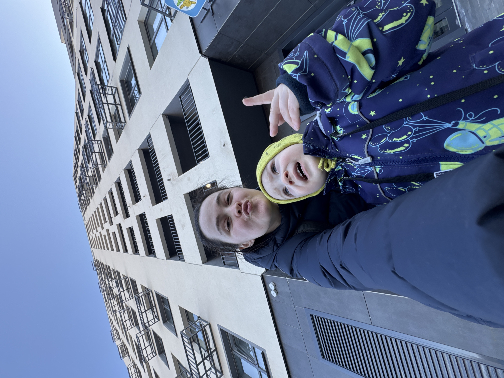 A selfie taken from a low angle shows a woman and a toddler standing outside a modern, high-rise building. The woman has dark hair pulled back and is making a kissing face at the camera. The toddler, wearing a yellow hat and a navy jacket patterned with balloons and stars, is smiling and pointing with his right hand. The building behind them is tall and slender with numerous windows and small balconies. The sky is visible above the building, suggesting a clear day. The bottom of the image shows a dark blue jacket and a gray concrete surface with metal grates. This image was captured in Izhevsk, Russia.