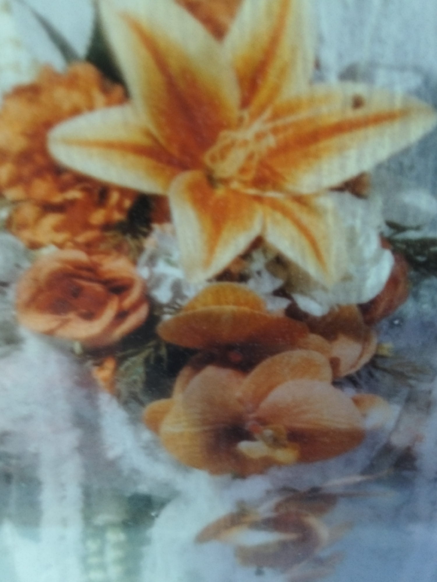 The image displays a close-up of an arrangement of flowers. The primary focus is a large, star-shaped lily with prominent orange and yellow stripes. To the left and below the lily are several smaller, peach-colored roses, some appearing more open than others. The background is a soft, light blue and white, suggesting a fabric or textured surface, possibly a tablecloth or a decorative filler. There are no people or discernible elements of an urban environment, such as buildings or landmarks, in the image. The floral arrangement appears to be indoors or in a setting where natural outdoor elements are not visible. The location of Cottrueng, Indonesia, is not evident from the visual information presented in the image.