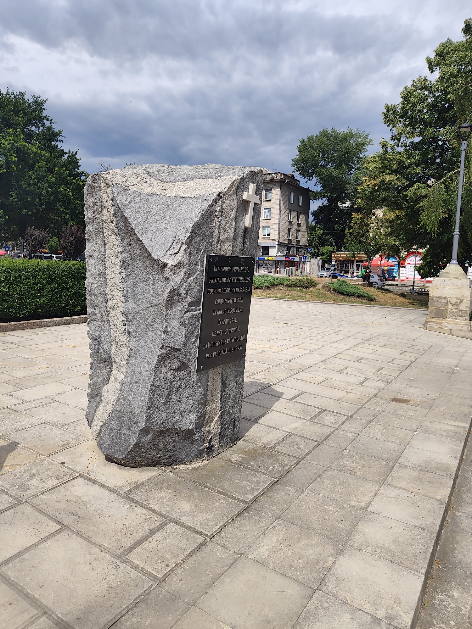 This is a frontal view of a monument located in Chișinău, Moldova. The monument is a large, rough-hewn grey stone with a metal plaque attached to its face. The plaque bears text in Romanian. A white cross is carved into the upper right portion of the stone. The monument is situated on a paved plaza with square stone tiles. In the background, there is a building with multiple stories, trees, and a lamppost. The sky is overcast with grey clouds. No people are clearly visible.