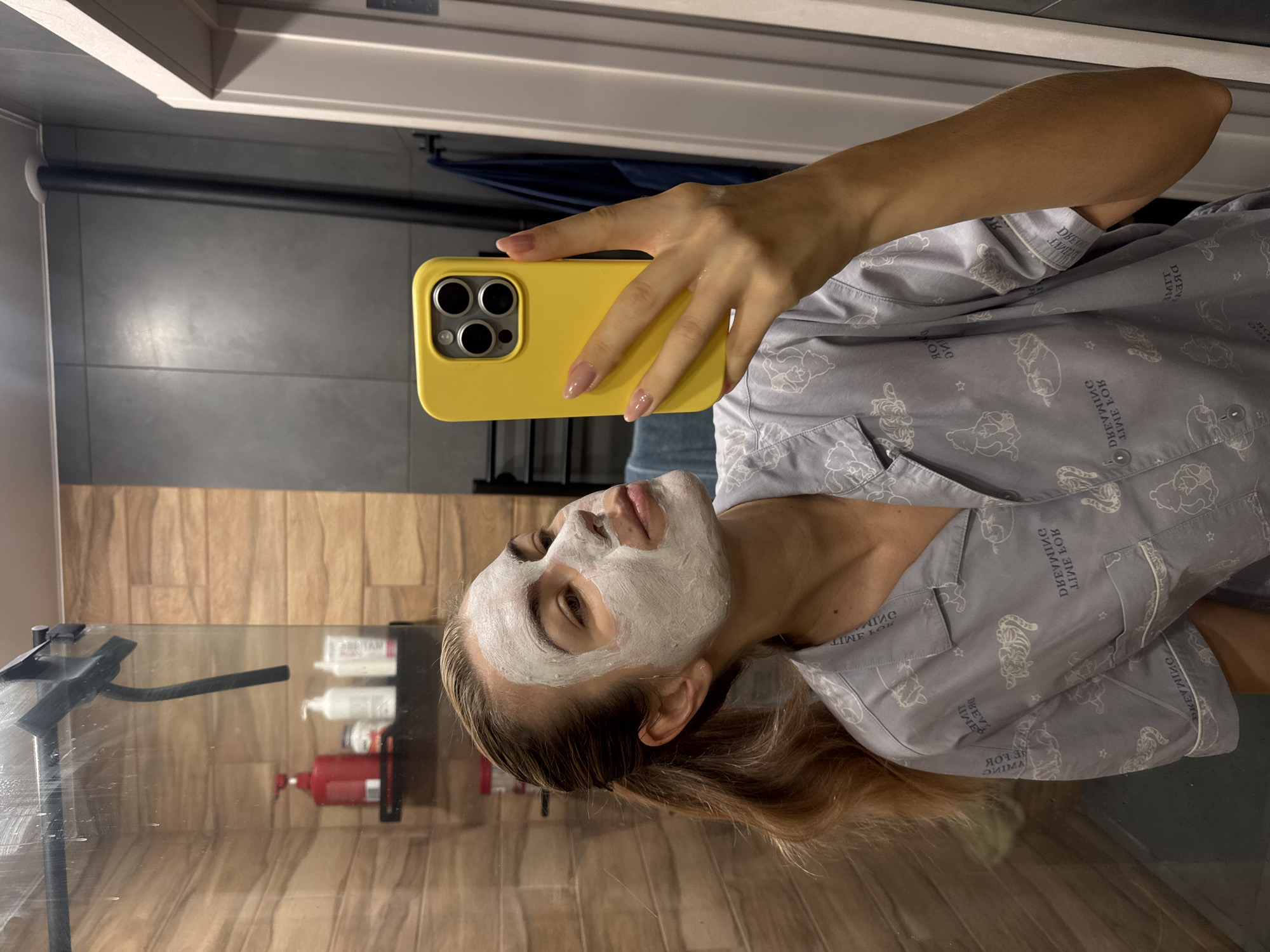 A person with a white facial mask is taking a selfie with a yellow smartphone in a bathroom. The individual is wearing patterned pajamas. The bathroom features a glass shower door and shelves with various bottles.