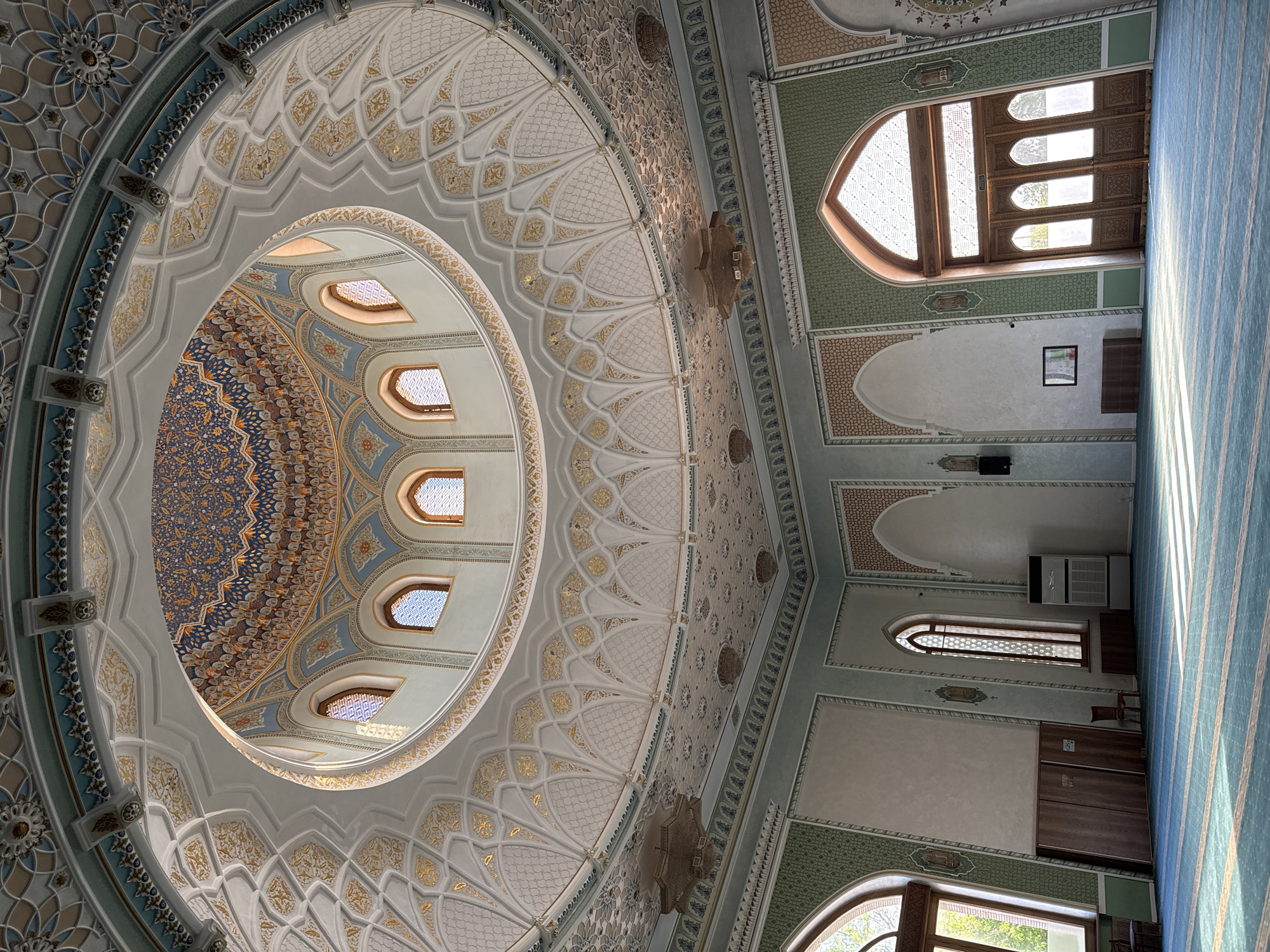 The image depicts the interior of a building, likely a mosque, in Tashkent, Uzbekistan. The primary focus is on the ceiling and a large dome-like structure with intricate geometric and floral patterns in gold, blue, and white. There are several small, arched windows around the inner circumference of the dome. The walls are decorated with similar patterns and archways. Natural light streams in through arched windows in the walls. A blue carpet with light stripes covers the floor. In the wall of the building, there is an artwork frame and a air conditioning unit. The perspective is looking upwards from below, emphasizing the height and detailed ornamentation of the architecture.