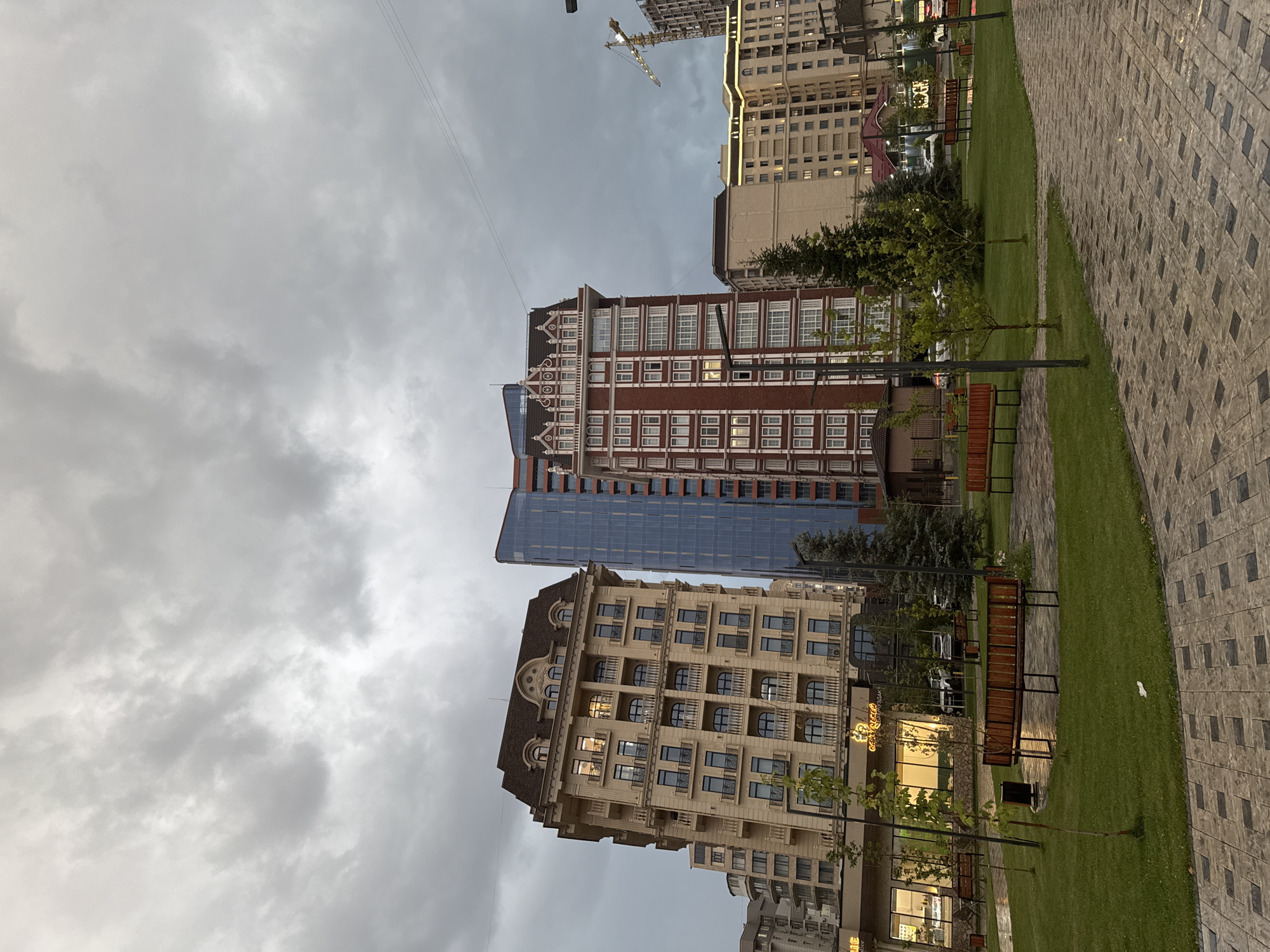 The image depicts an overcast day in a modern urban park area, likely in Bishkek, Kyrgyzstan, given the architectural style and construction cranes indicative of development. The foreground features a paved walkway with a patterned design, leading into a well-maintained grassy area. Several wooden park benches are strategically placed throughout the green space, some appearing wet, suggesting recent rain. Small trees are scattered across the lawn.
Dominating the background are several multi-story buildings. On the left, a light-colored building with a mansard roof and numerous balconies is prominent. To its right stands a sleek, modern high-rise with a glass facade in shades of blue and silver. Adjacent to this is a taller, brick-colored building with ornate white trim and numerous windows. Further in the distance, more apartment complexes and construction sites are visible, including a yellow construction crane. The sky is heavily clouded with a neutral, gray tone, and no people are visible in the scene. The lighting is diffused, creating a somewhat subdued atmosphere.