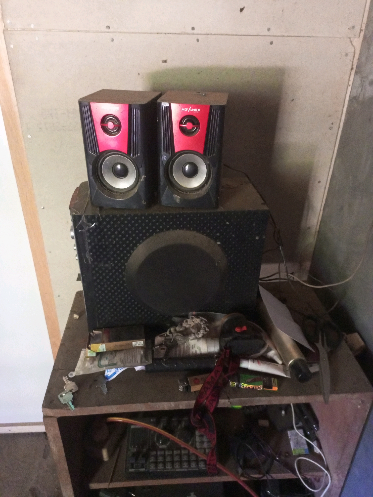 In Jatipurno, Indonesia, a medium shot captures a rustic, cluttered wooden shelf unit. The unit holds a black subwoofer with a patterned grill, topped by two black and red satellite speakers, each featuring a silver speaker cone and the "ADVANCE" logo. Scattered across the shelf are various items including keys, a lighter, cigarette packs, scissors, and what appears to be a gold-colored cylinder. Cables are visible, some organized and others loose, connecting to electronic components housed within the lower section of the shelf unit. The background consists of textured, light-colored walls and a rough concrete wall on the right, suggesting an unfinished or industrial setting. The lighting is somewhat dim, casting shadows that add to the scene's raw, unpolished feel.