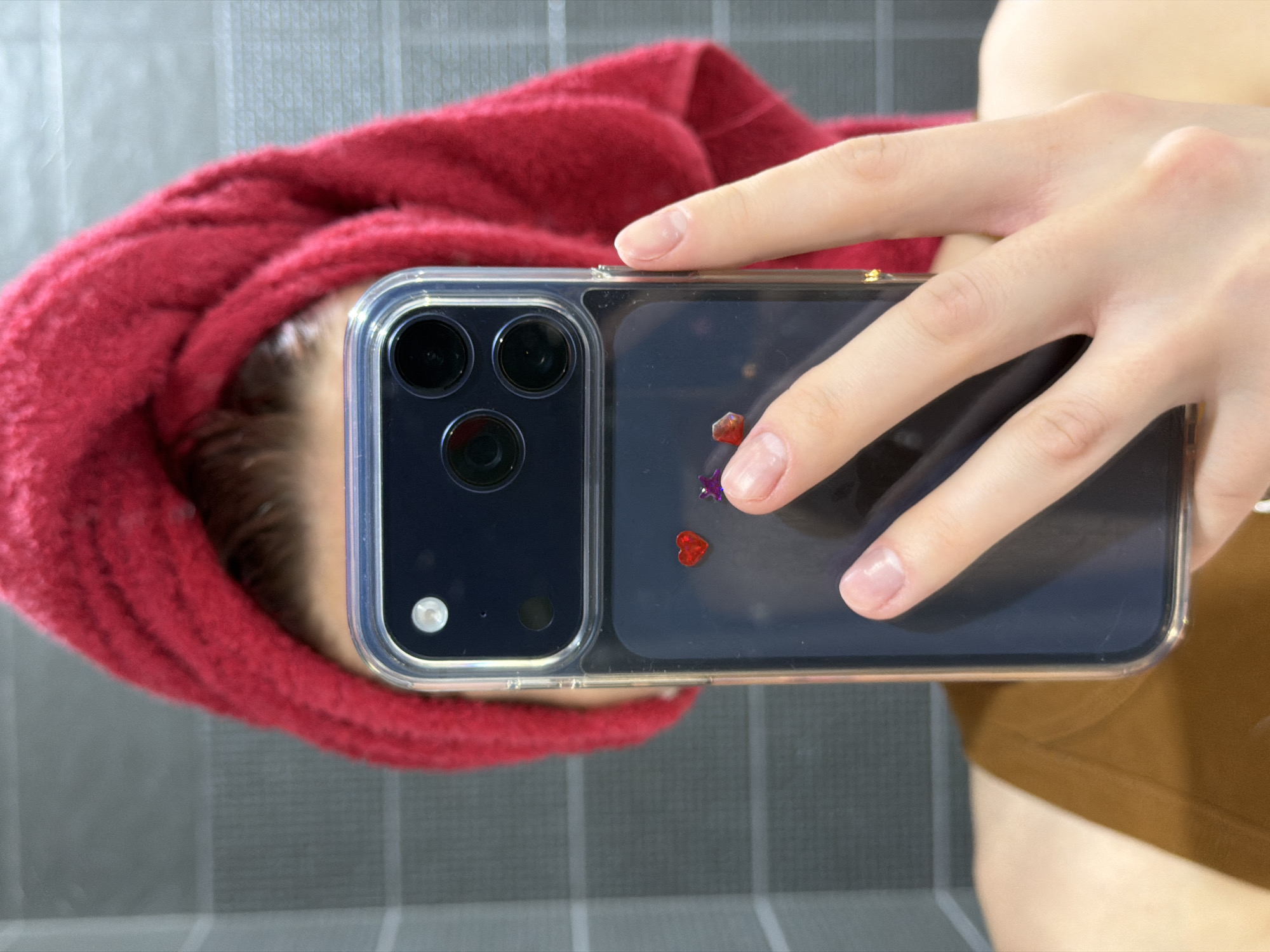 A person is holding a cell phone up to a mirror, obscuring their face. A red towel is wrapped around their head. The cell phone is in a clear case with glittery decorations. The person is wearing a brown top. The background appears to be a gray tiled wall. The location and city of Chaikovsky, Russia, cannot be determined from the image.