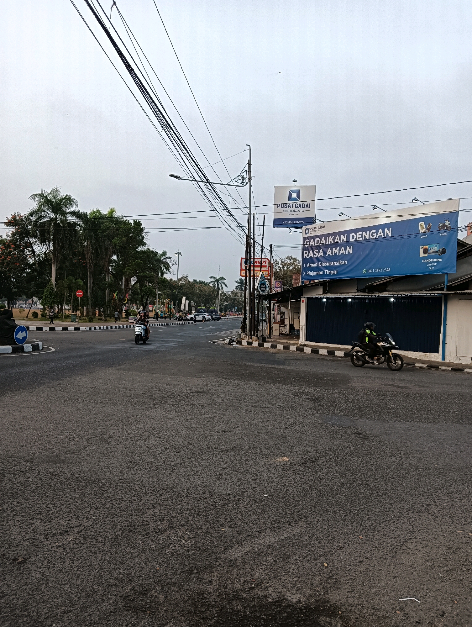 The image shows a street intersection in Purworejo, Indonesia, under an overcast sky. In the foreground, the asphalt road surface is visible, with some wet patches. Two motorcycles are present: one white scooter with a rider is moving through the intersection, and a black and yellow motorcycle with a rider is proceeding along the side of the road.
To the left of the intersection, a round traffic island is marked with black and white stripes and features a blue directional arrow sign pointing left. Beyond the island, there is a sidewalk with pedestrians and lush green palm trees and other foliage. A red circular traffic sign with a horizontal bar is also visible on the left.
In the background, several buildings and billboards are present. A prominent blue billboard reads "Gadaikan Dengan Rasa Aman" (Pledge with Peace of Mind) and lists services like "Aman Diasuransikan" (Secured and Insured) and "Pinjaman Tinggi" (High Loan). Smaller signs indicate "Pusat Gadai Indonesia" (Pawnshop Indonesia). Another billboard depicts items like gold, a laptop, a smartphone, and a DSLR camera, along with the text "Perhiasan, Emas, BPKB, Handphone, Laptop, SLR." A street light is visible. Further in the distance, more trees and buildings are scattered, and several cars are on the road. A small bird is visible in the sky. The overall environment appears to be an urban setting with a mix of commercial establishments and green spaces.