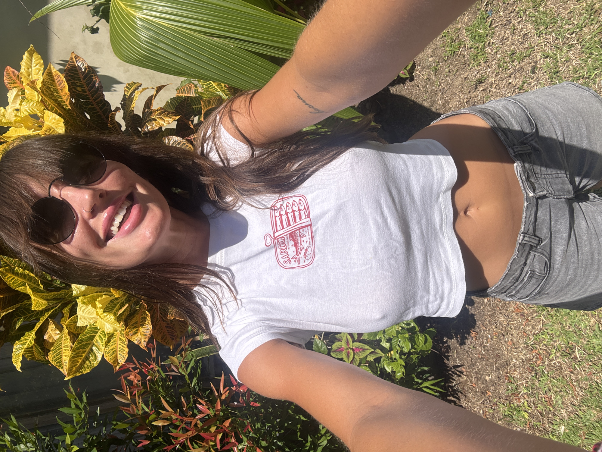 A young woman with long brown hair and sunglasses is taking a selfie outdoors in North Kuta, Indonesia. She is wearing a white cropped t-shirt with a red sardine can graphic and gray jeans. Her arms are visible, with a tattoo on one. She is surrounded by tropical plants, including yellow and green foliage. The ground appears to be a mix of grass and dirt.