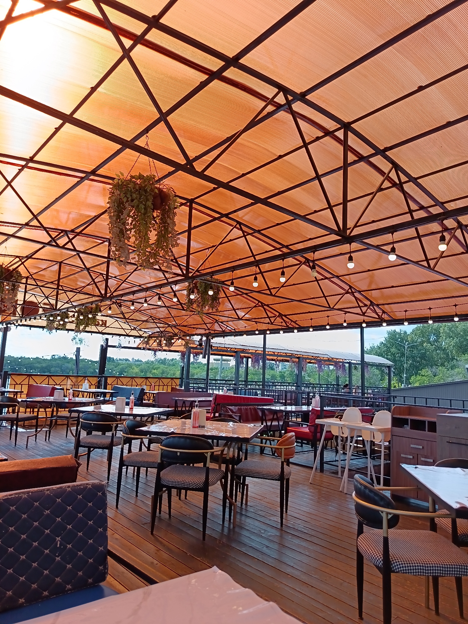 This is an exterior shot of a restaurant with an orange canopy and wooden deck. The canopy is supported by a dark metal frame and illuminated by string lights. Several tables and chairs are set up on the deck, some with red seating. In the background, there is a view of trees and a body of water, suggesting a location in or near Qarağandı city administration, Kazakhstan. There are no people visible in the image.