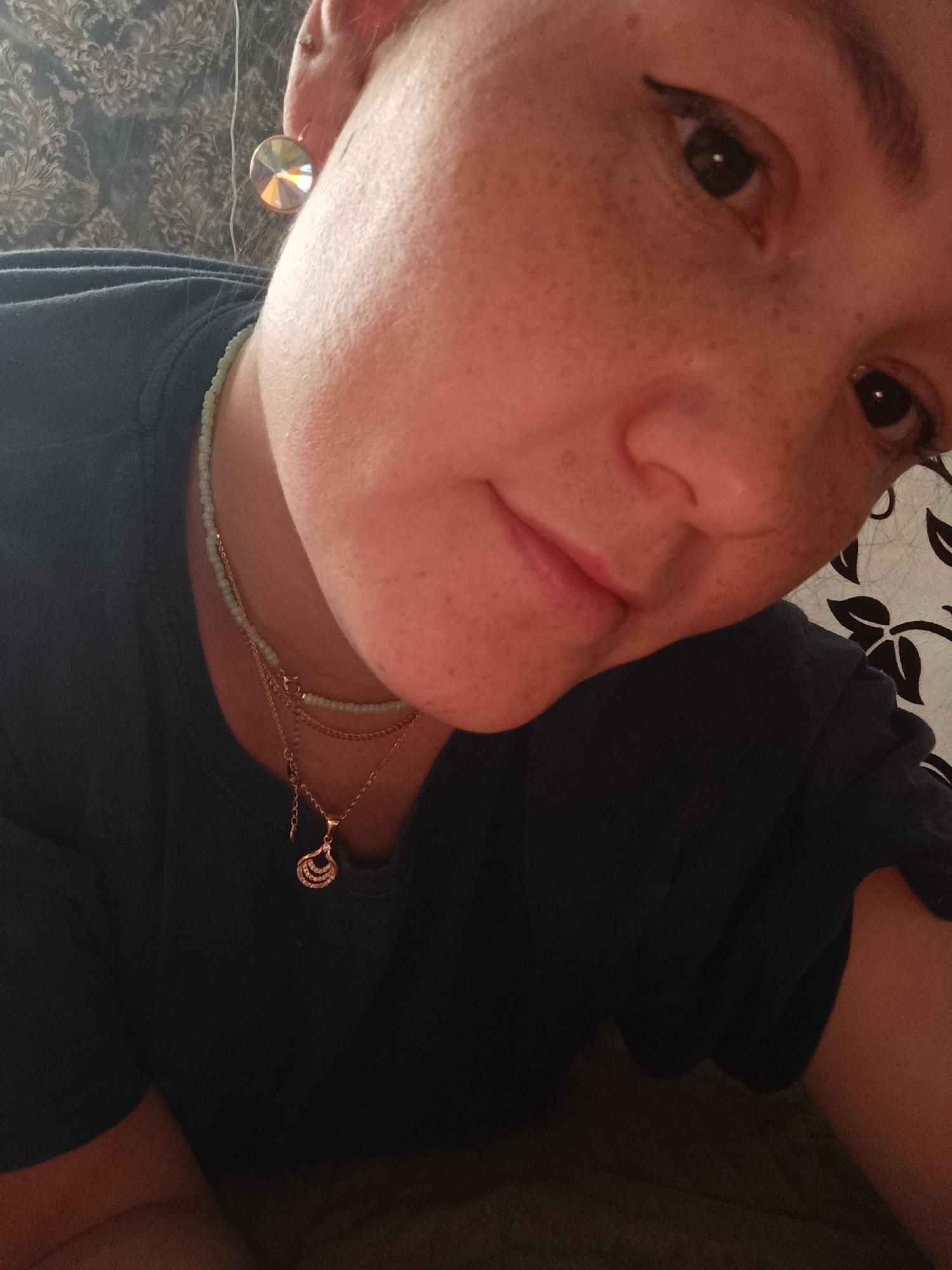This is a close-up, indoor, head and shoulder portrait of a young woman with fair skin and visible freckles. She is wearing a dark blue t-shirt, a light green beaded necklace, and a delicate rose gold layered necklace with a teardrop-shaped pendant adorned with small clear stones. She also has a large, iridescent, faceted earring in her left ear. Her eyes are dark and appear to have eyeliner applied. The background consists of a textured wallpaper with a floral pattern and a dark fabric. The lighting suggests it is artificial. There is no discernible location within the city of Karsha, Russia.