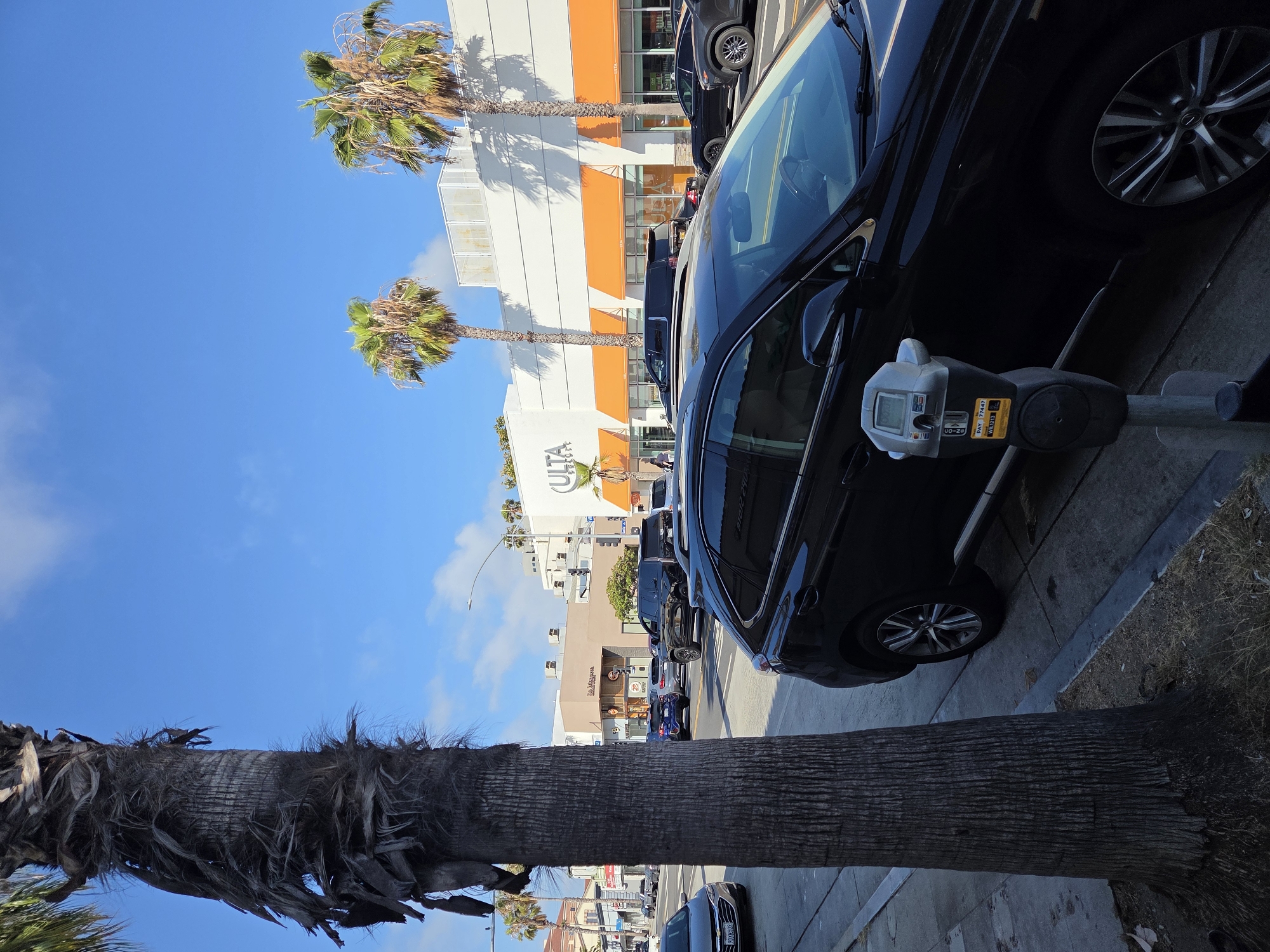The image depicts a street scene in Santa Monica, California, USA. In the foreground, a large palm tree trunk stands to the left, partially obscuring the view. A black SUV is parked at the curb, with a parking meter in front of it. The meter is silver and gray with a digital display and payment instructions. Behind the SUV, more cars are visible on the street, suggesting moderate traffic. In the background, a white building with orange awnings houses a store labeled "ULTA BEAUTY." Several palm trees are planted along the sidewalk in front of the building, and the sky is blue with scattered clouds. The overall impression is a sunny day in a commercial area of the city.